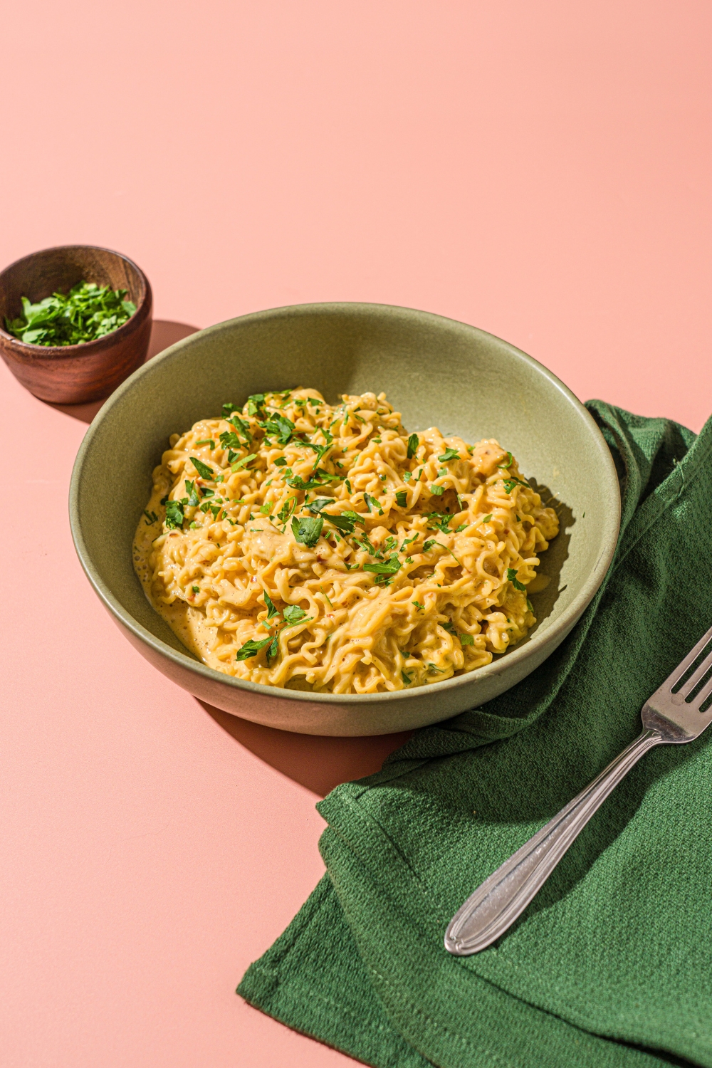 A green bowl with cheesy Maggi noodles garnished with fresh cilantro. The bowl is on a pink counter with a green cloth napkin, fork, and small bowl of garnishes.
