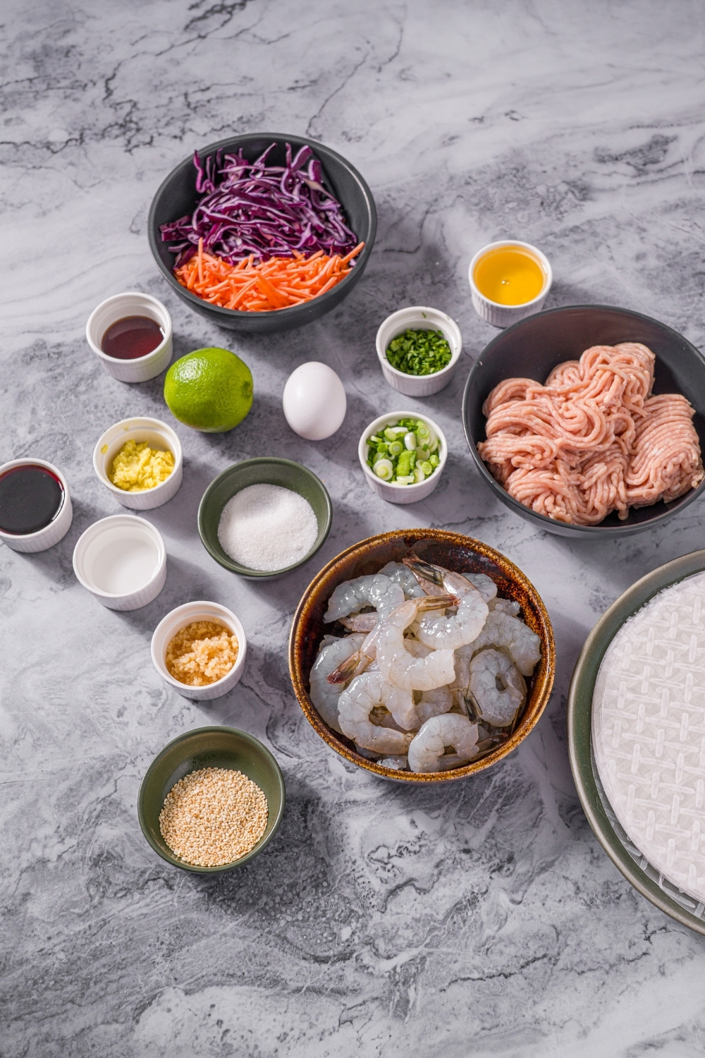 A marble counter with bowls in various sizes containing ingredients to make chicken and shrimp rice paper dumplings including rice paper wrappers, ground chicken, shrimp, shredded carrots, shredded cabbage, onions, ginger, garlic, egg, sesame oil, soy sauce, fish sauce, and more.