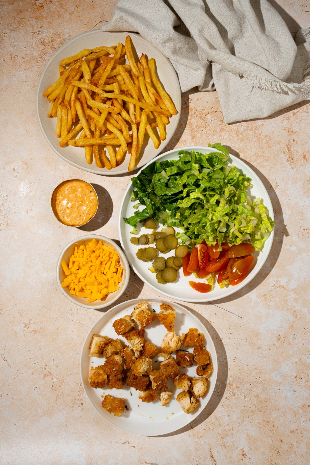 An overhead shot of several plates in various sizes containing ingredients to make chicken burger bowl loaded fries including fries, crispy chicken, shredded cheese, lettuce, pickles, tomatoes, and burger sauce.
