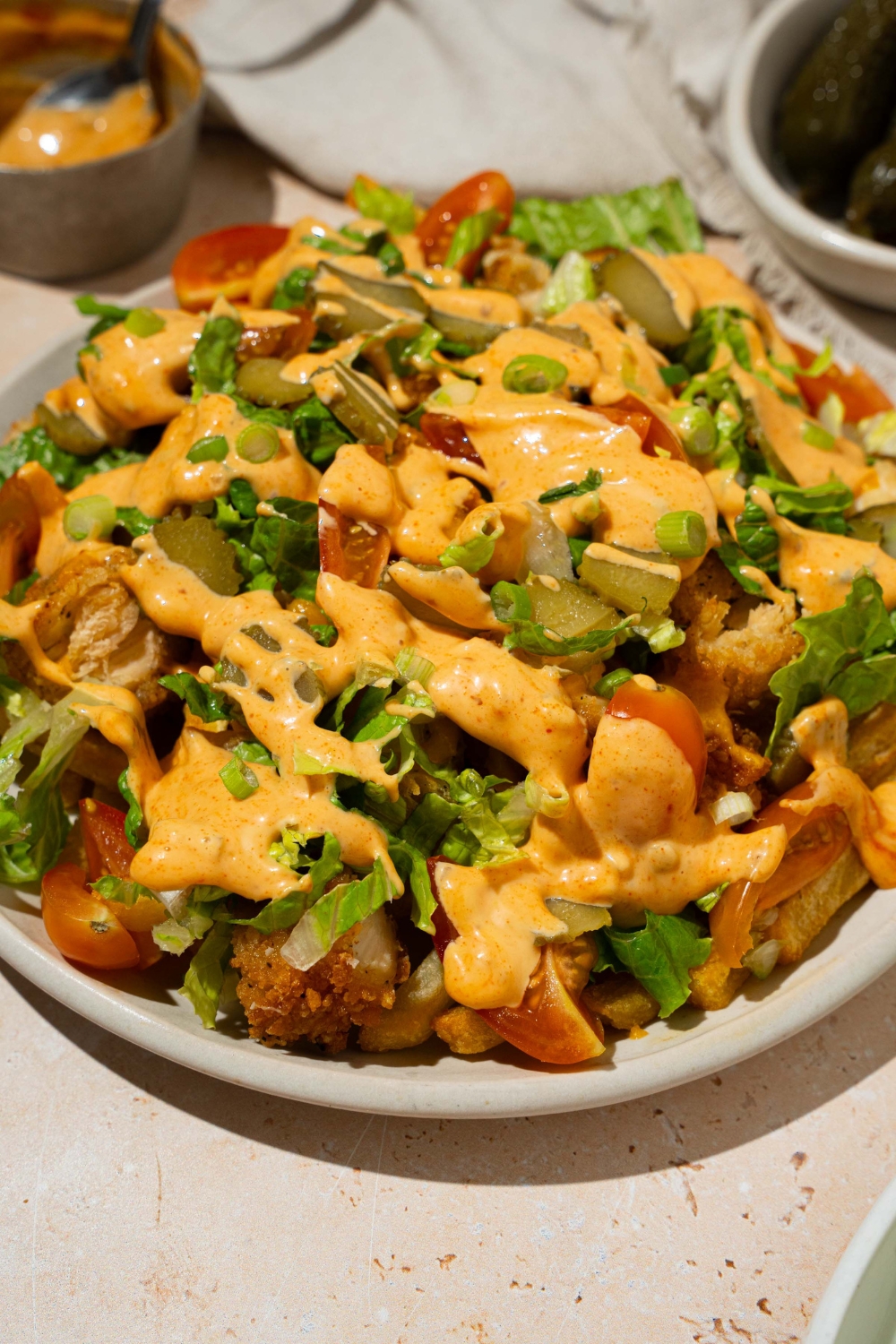 A white plate with chicken burger bowl loaded fries with fries topped with crispy chicken, lettuce, shredded cheese, tomatoes, and pickles drizzled with burger sauce. The plate is on a tan counter with a white cloth napkin and small bowl of burger sauce.