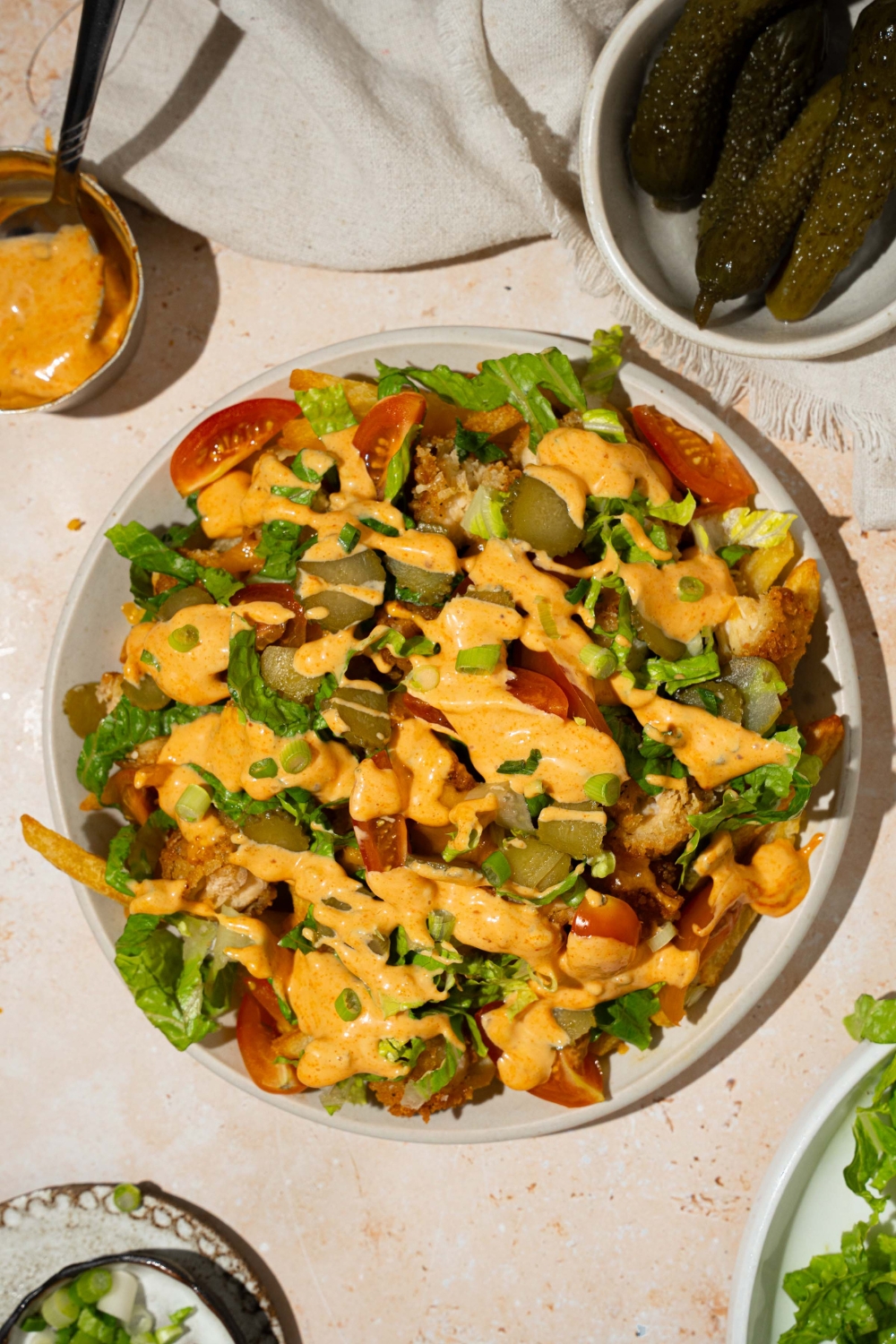A white plate with chicken burger bowl loaded fries with fries topped with crispy chicken, lettuce, shredded cheese, tomatoes, and pickles drizzled with burger sauce. The plate is on a tan counter with a white cloth napkin, small bowl of pickles, and small bowl of burger sauce.