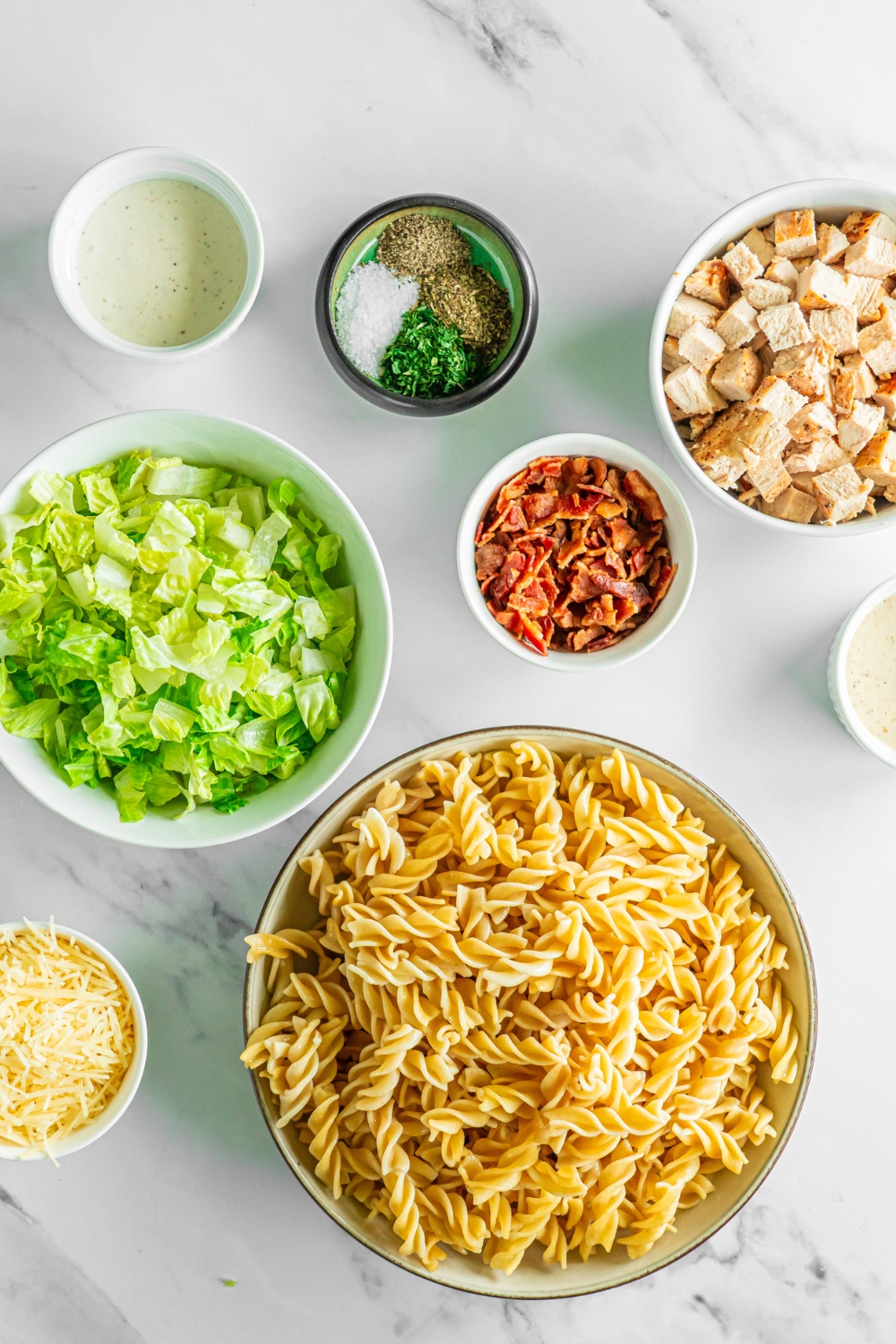 A marble counter with several bowls in various sizes containing ingredients to make chicken caesar pasta salad including rotini pasta, shredded lettuce, bacon, cubed chicken, shredded parmesan cheese, caesar dressing, and seasonings.