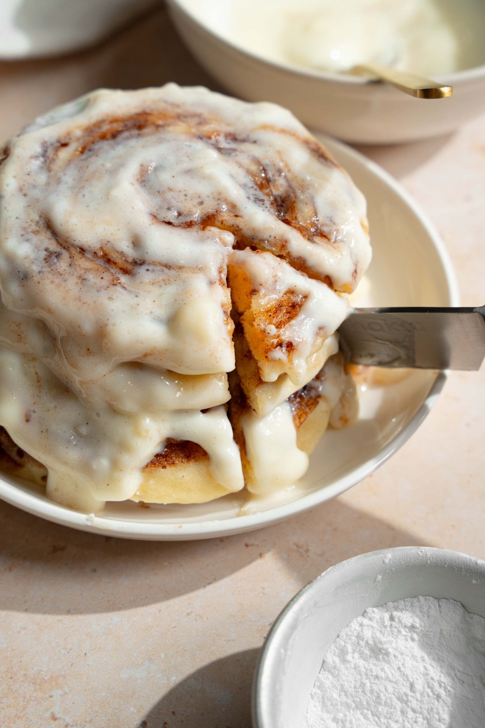 A plate with a stack of cinnamon roll pancakes topped with cream cheese icing. There is a knife taking a bite from the stack. The plate is on a tan counter with a bowl of cream cheese icing.