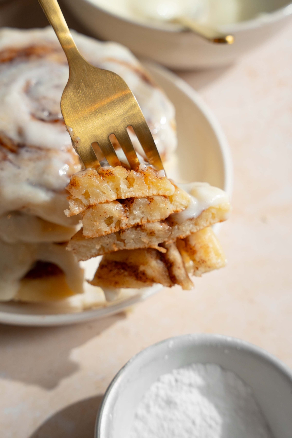 A close up of a fork with a bite of cinnamon roll pancakes. There is a plate with a stack of pancakes on a tan counter blurred in the background.