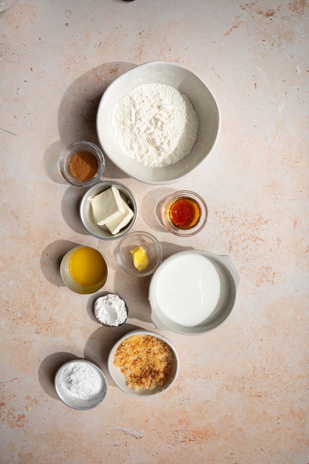 An overhead shot of several bowls in various sizes containing ingredients to make cinnamon roll pancakes including flour, cinnamon, butter, vanilla, powdered sugar, brown sugar, vanilla, and maple syrup.