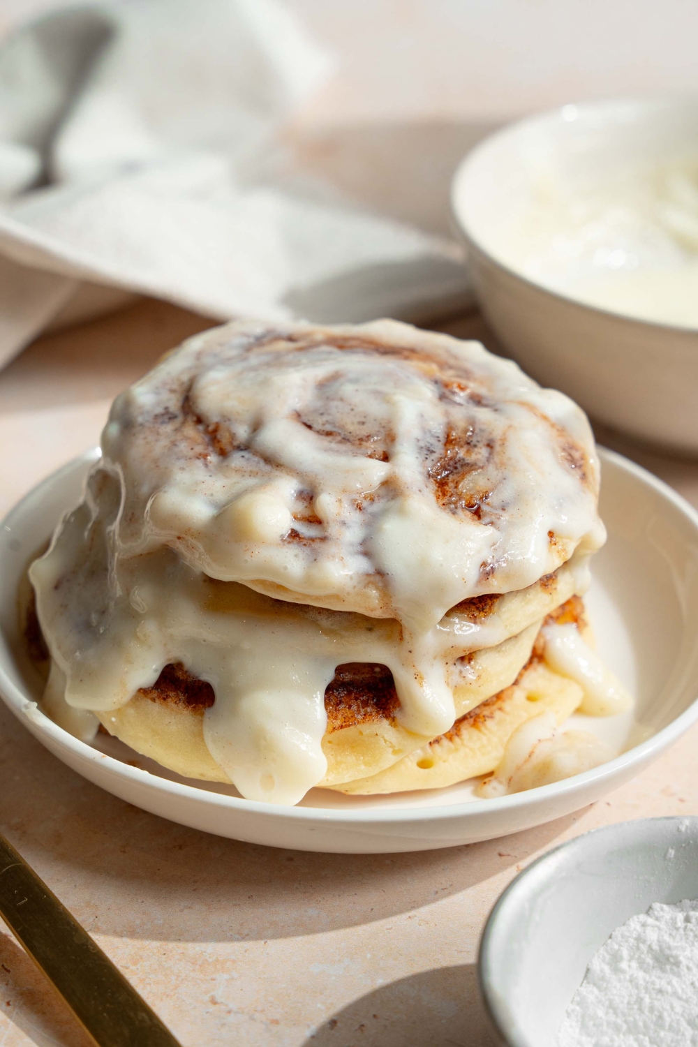 A white plate with a stack of cinnamon roll pancakes topped with cream cheese icing. The plate is on a tan counter with a bowl of cream cheese icing.