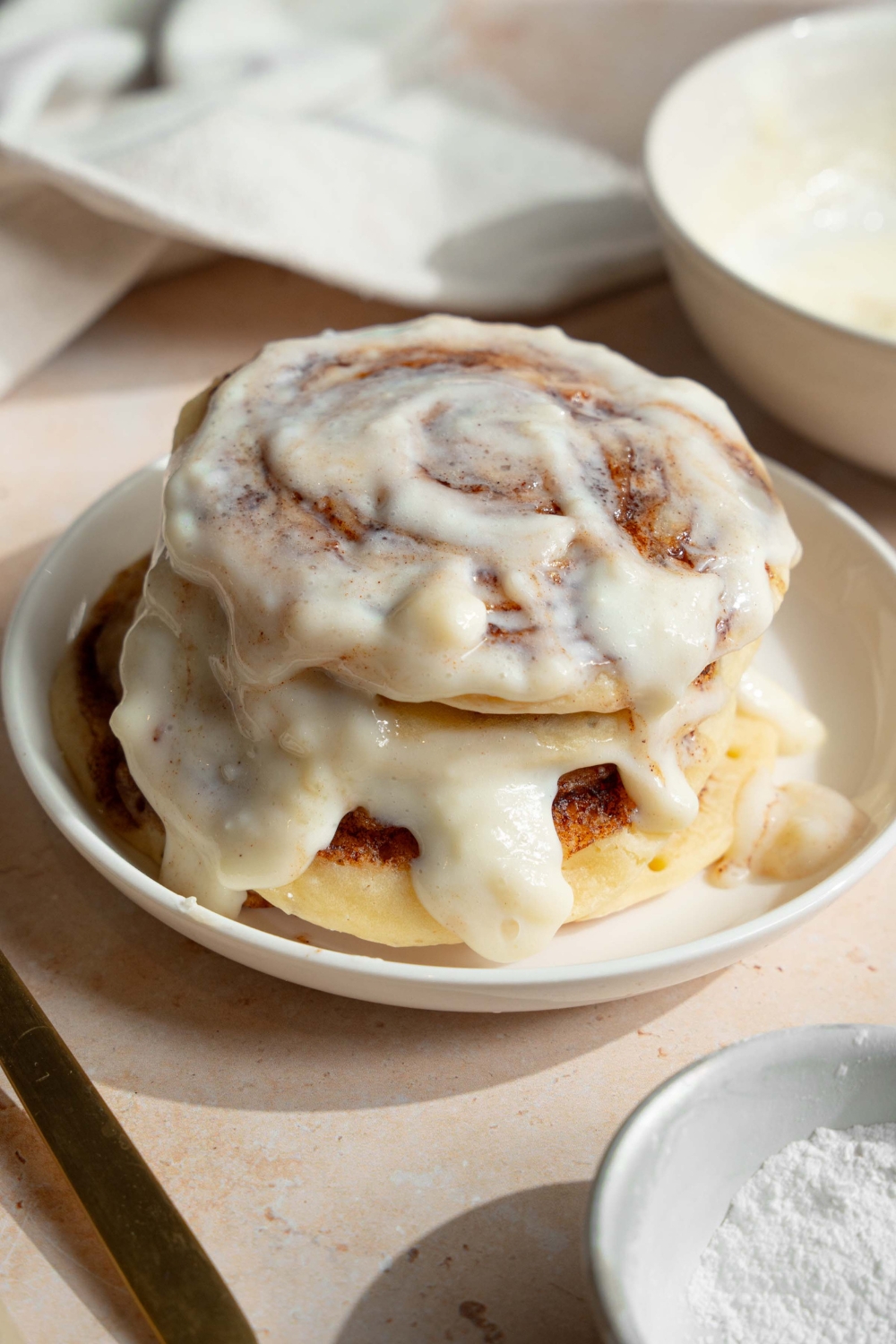 A white plate with a stack of cinnamon roll pancakes topped with cream cheese icing. The plate is on a tan counter with a bowl of cream cheese icing.