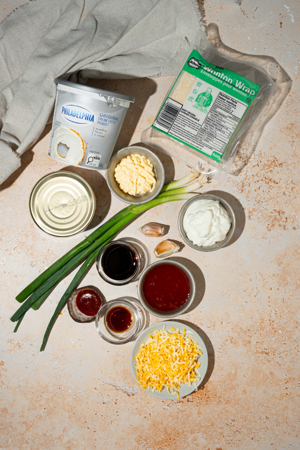 An overhead shot of several bowls in various sizes containing ingredients to make crab rangoon dip including cream cheese, crab meat, mayonnaise, sour cream, garlic, green onions, soy sauce, shredded cheese, and wonton wrappers.