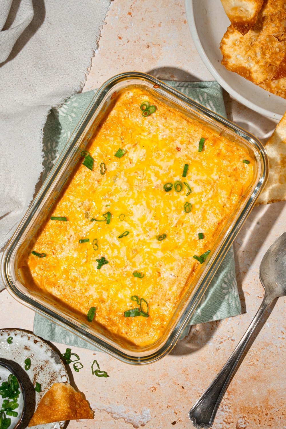A glass baking dish with baked crab rangoon dip garnished with sliced green onions. The dish is on a tan counter with a bowl of wonton chips, white cloth napkin, and spoon.