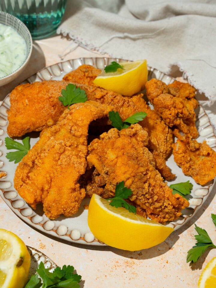 A white plate with several pieces of crispy Southern style fried fish garnished with fresh parsley and served with lemon. The plate is on a tan counter with a white cloth napkin.