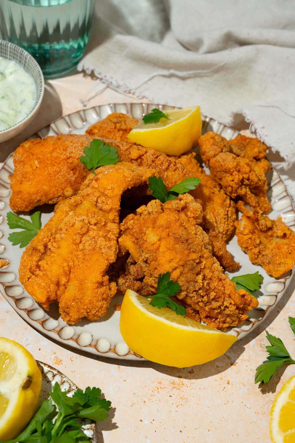 A white plate with several pieces of crispy Southern style fried fish garnished with fresh parsley and served with lemon. The plate is on a tan counter with a white cloth napkin.