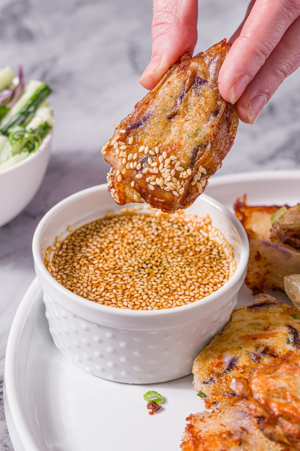 A white plate with a hand dipping a chicken and shrimp rice paper dumpling into a small bowl of dipping sauce with sesame seeds. There are additional dumplings on the plate. The plate is on a marble counter.