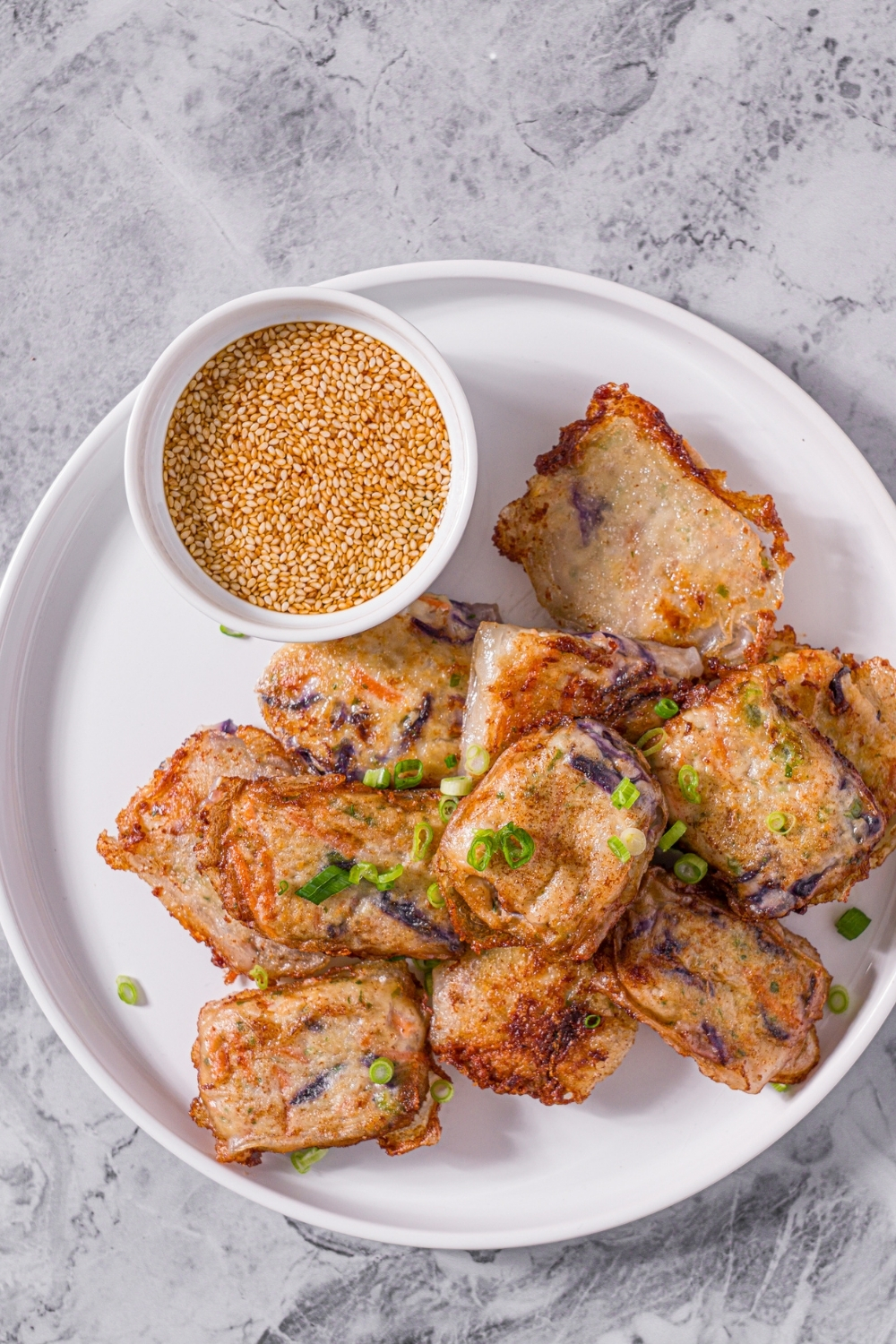A white plate with chicken and shrimp rice paper dumplings garnished with sliced green onions and served with a small bowl of dipping sauce. The plate is on a marble counter.