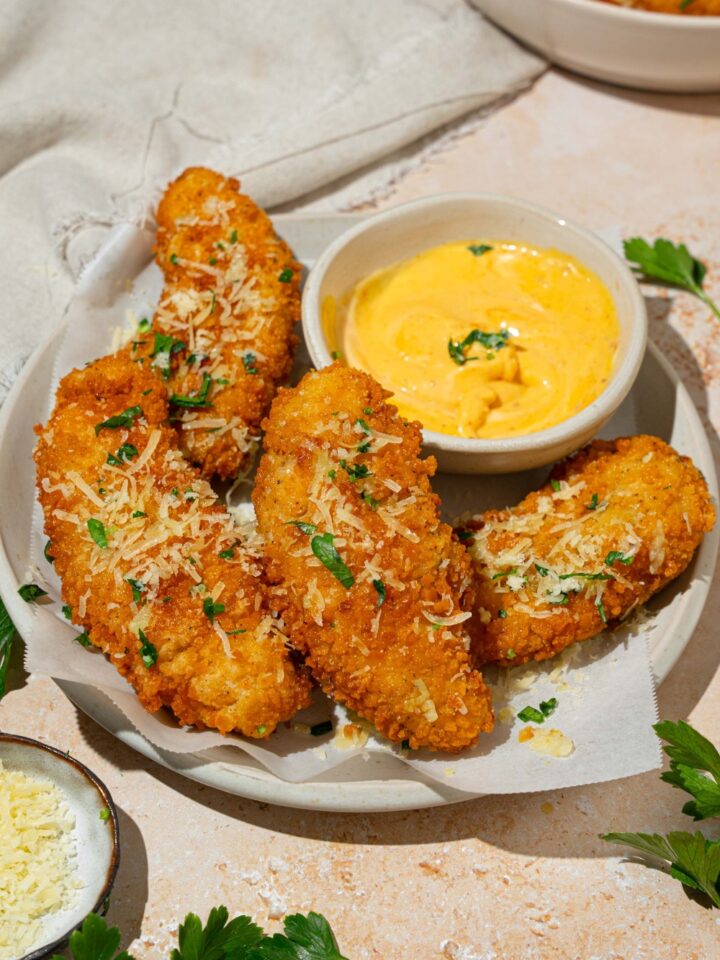 A white plate lined with parchment paper with garlic parmesan chicken tenders garnished with parmesan cheese and fresh parsley. There is a bowl of dip on the plate. The plate is on a tan counter with a white cloth napkin and small bowl of cheese.