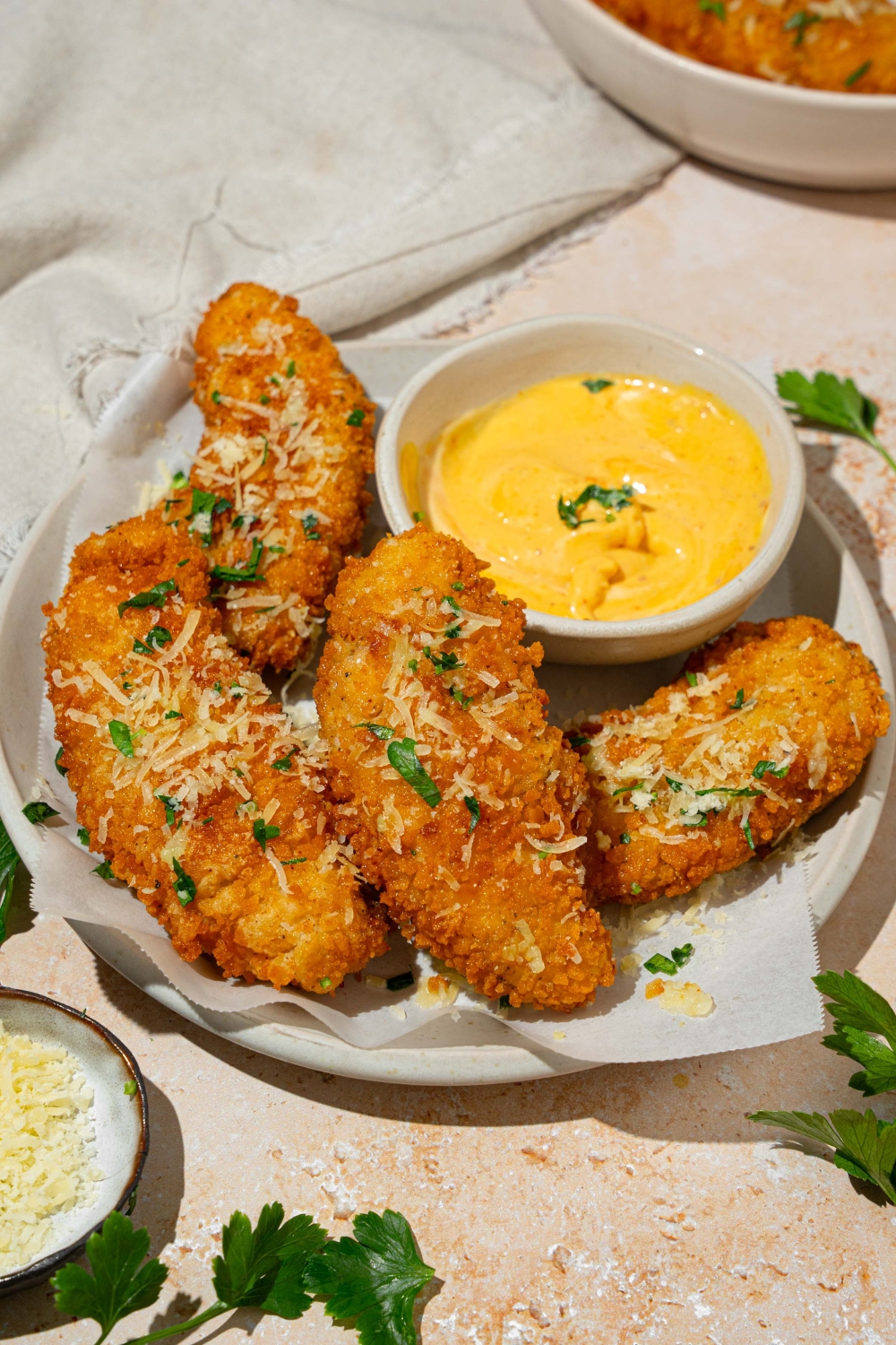 A white plate lined with parchment paper with garlic parmesan chicken tenders garnished with parmesan cheese and fresh parsley. There is a bowl of dip on the plate. The plate is on a tan counter with a white cloth napkin and small bowl of cheese.
