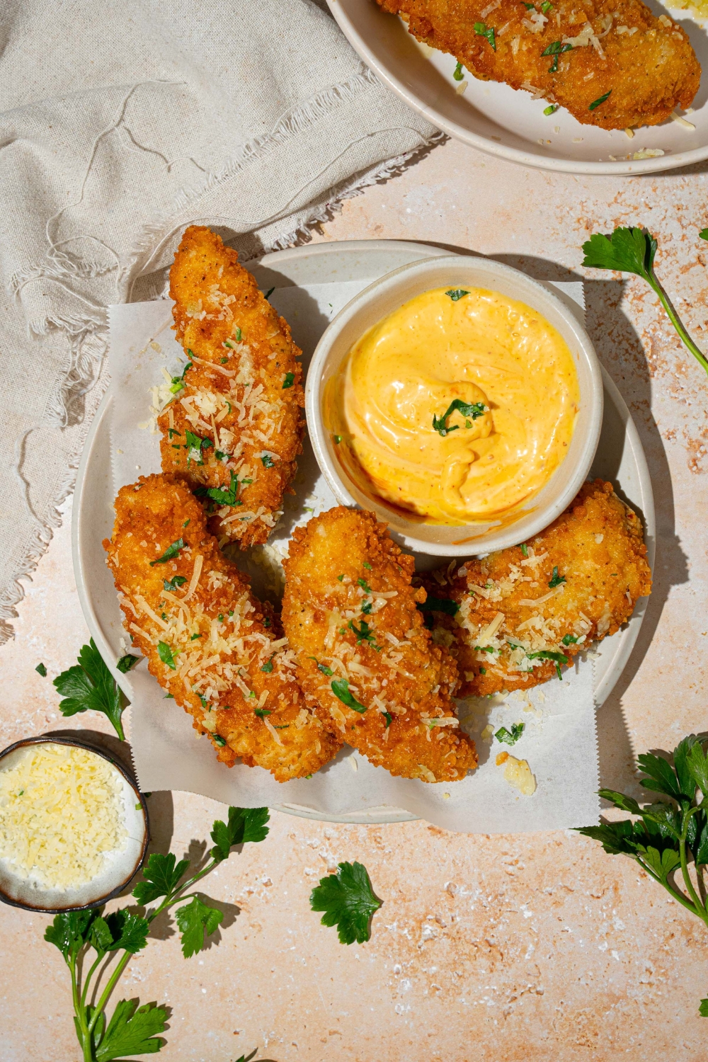 A white plate lined with parchment paper with garlic parmesan chicken tenders garnished with parmesan cheese and fresh parsley. There is a bowl of dip on the plate. The plate is on a tan counter with a white cloth napkin and small bowl of cheese.