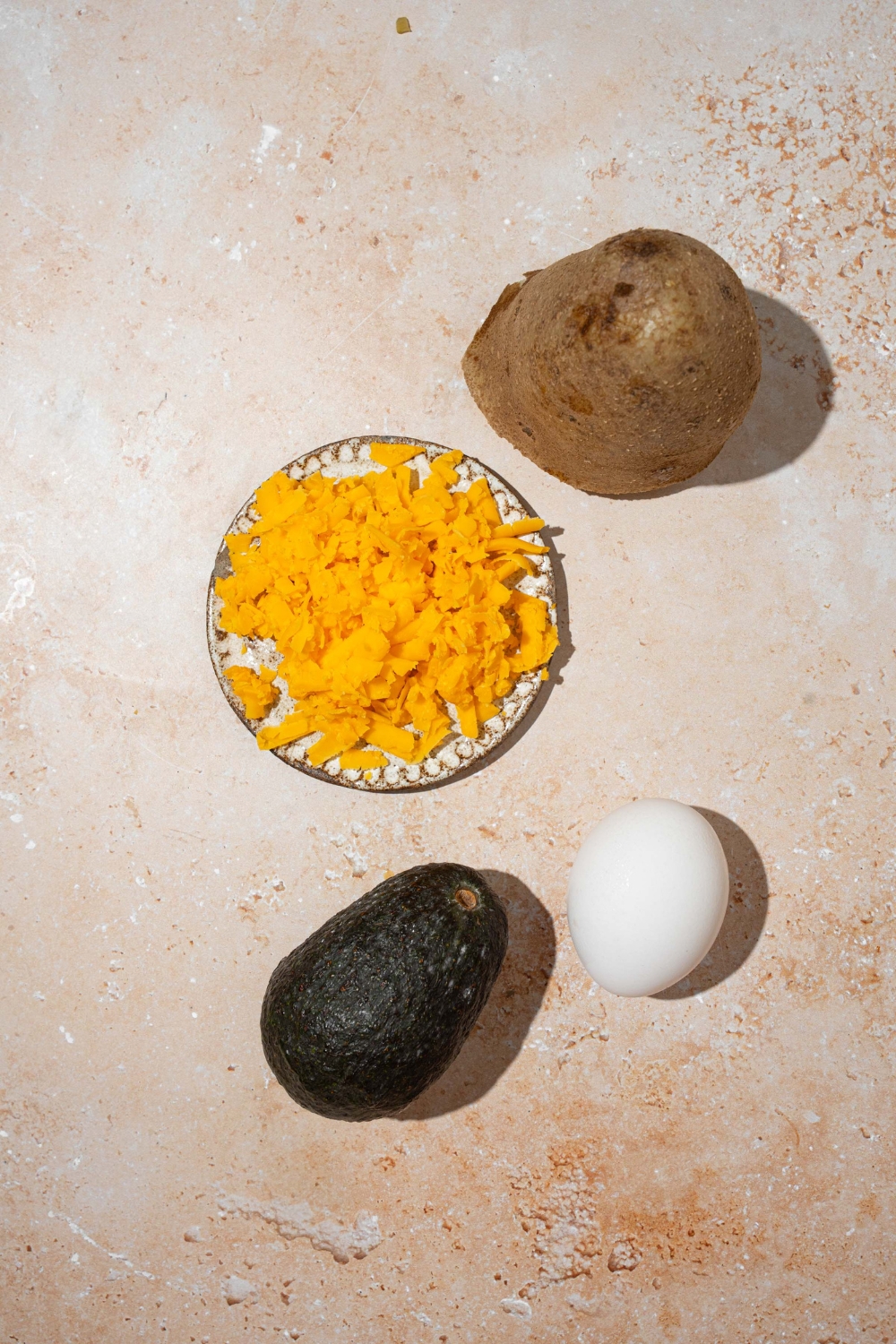 A tan counter with ingredients to make crispy potato waffles including shredded cheese, a potato, an egg, and an avocado.