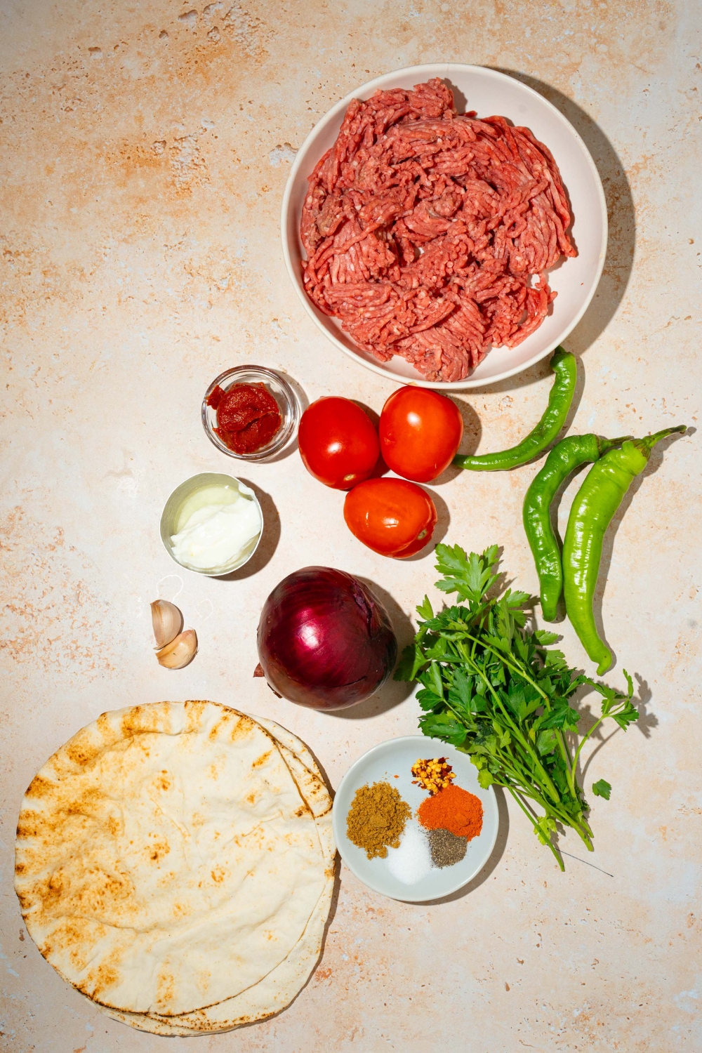A tan counter with ingredients to make doner kebab including ground beef, onion, tomatoes, greek yogurt, tomato paste, green peppers, fresh parsley, garlic, pita bread, and seasonings.