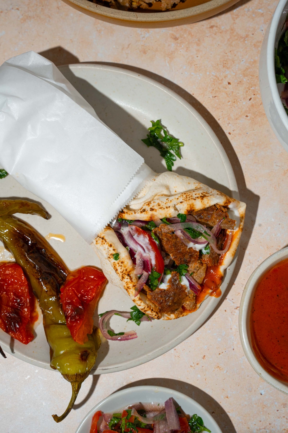 A white plate with a doner kebab wrapped in pita bread with garlic sauce and roasted vegetables with a yogurt sauce. The pita is wrapped in paper. The wrap is on a white plate with roasted green peppers and roasted tomatoes. The plate is on a tan counter.