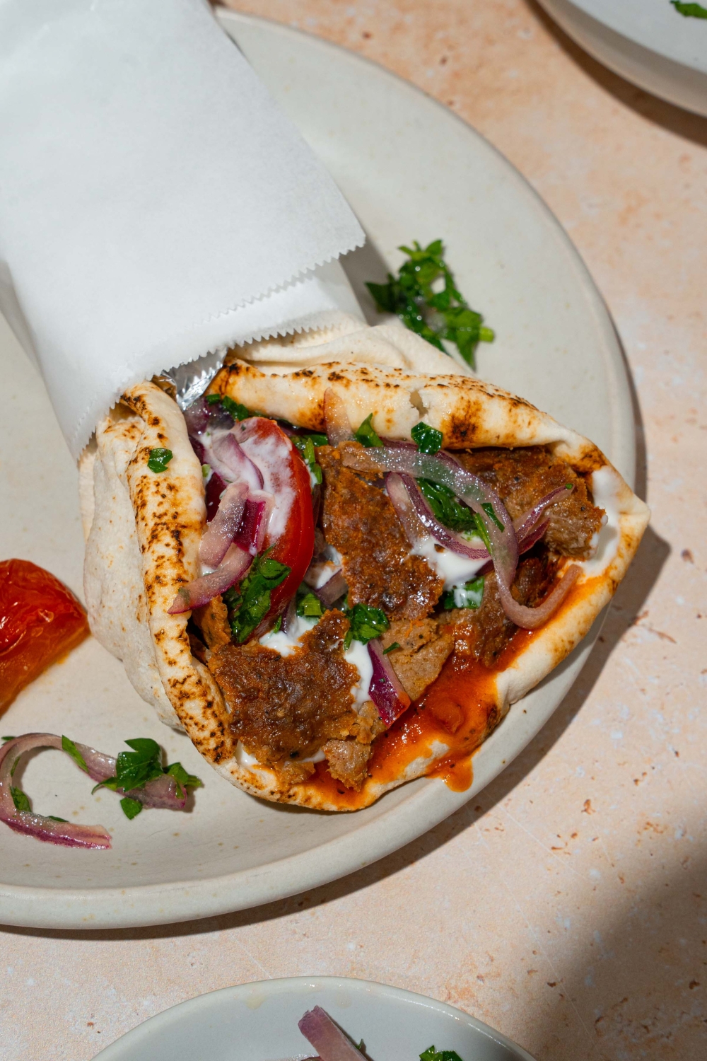 A white plate with a doner kebab wrapped in pita bread with garlic sauce and roasted vegetables with a yogurt sauce. The pita is wrapped in paper. The wrap is on a white plate with roasted green peppers and roasted tomatoes. The plate is on a tan counter.