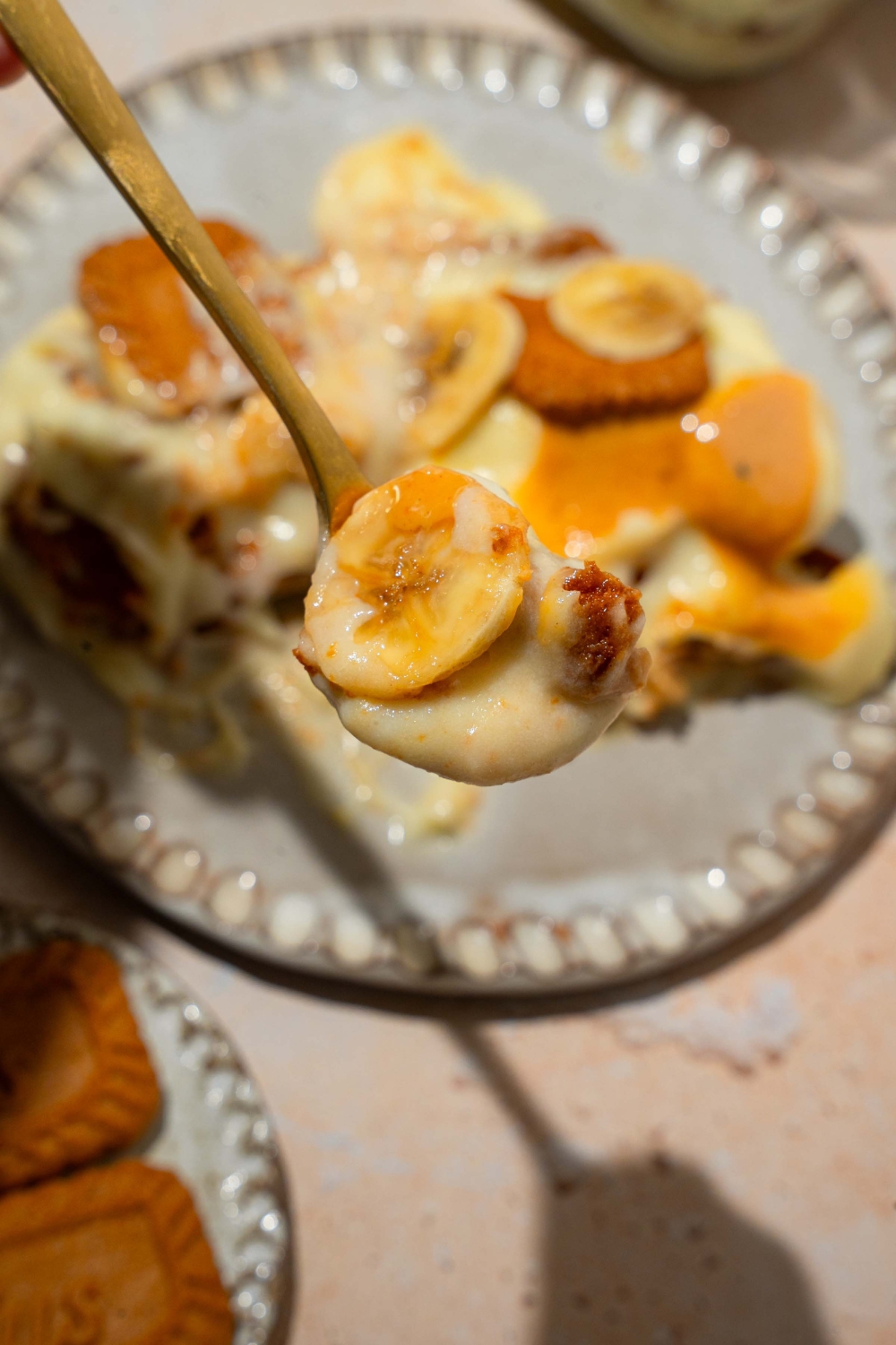 A close up of a spoon with a bite of Biscoff banana pudding with a plte of pudding blurred in the background. The plate is on a tan counter.