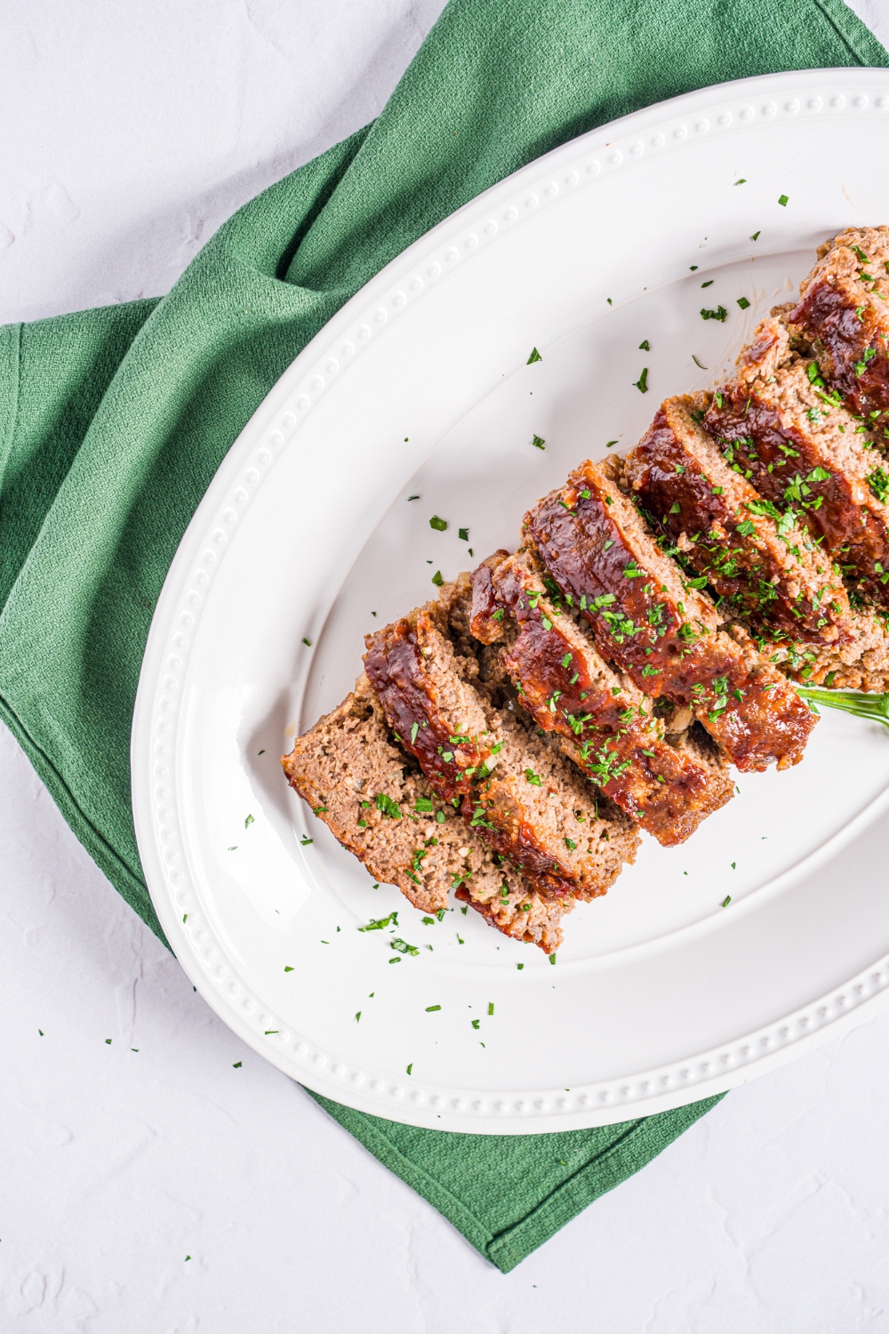 A white platter with sliced meatloaf without eggs. The meatloaf is garnished with fresh parsley. The plate is on a marble counter with a green cloth napkin.