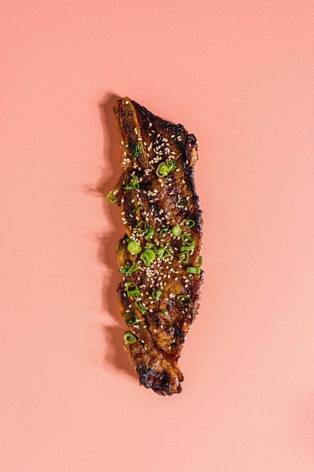 A strip of flanken style short rib garnished with sesame seeds and sliced green onion. The rib is on a pink counter.