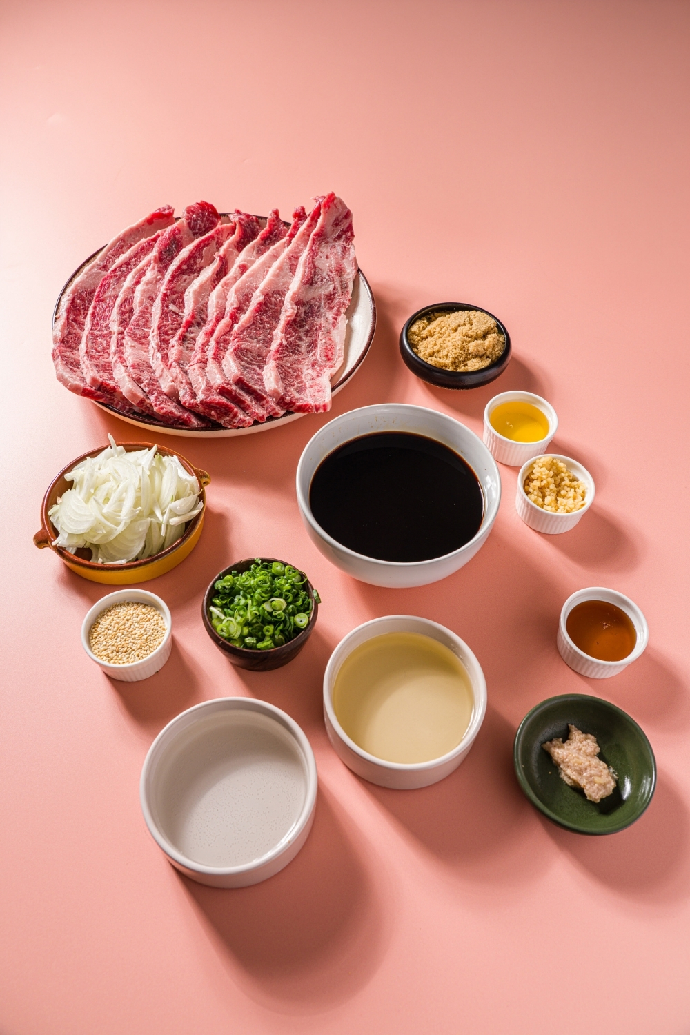 Several bowls in various sizes containing ingredients to make flanken style short ribs including flanken beef short ribs, soy sauce, mirin, brown sugar, honey, ginger, garlic, sliced onion, sliced green onion, and sesame oil.