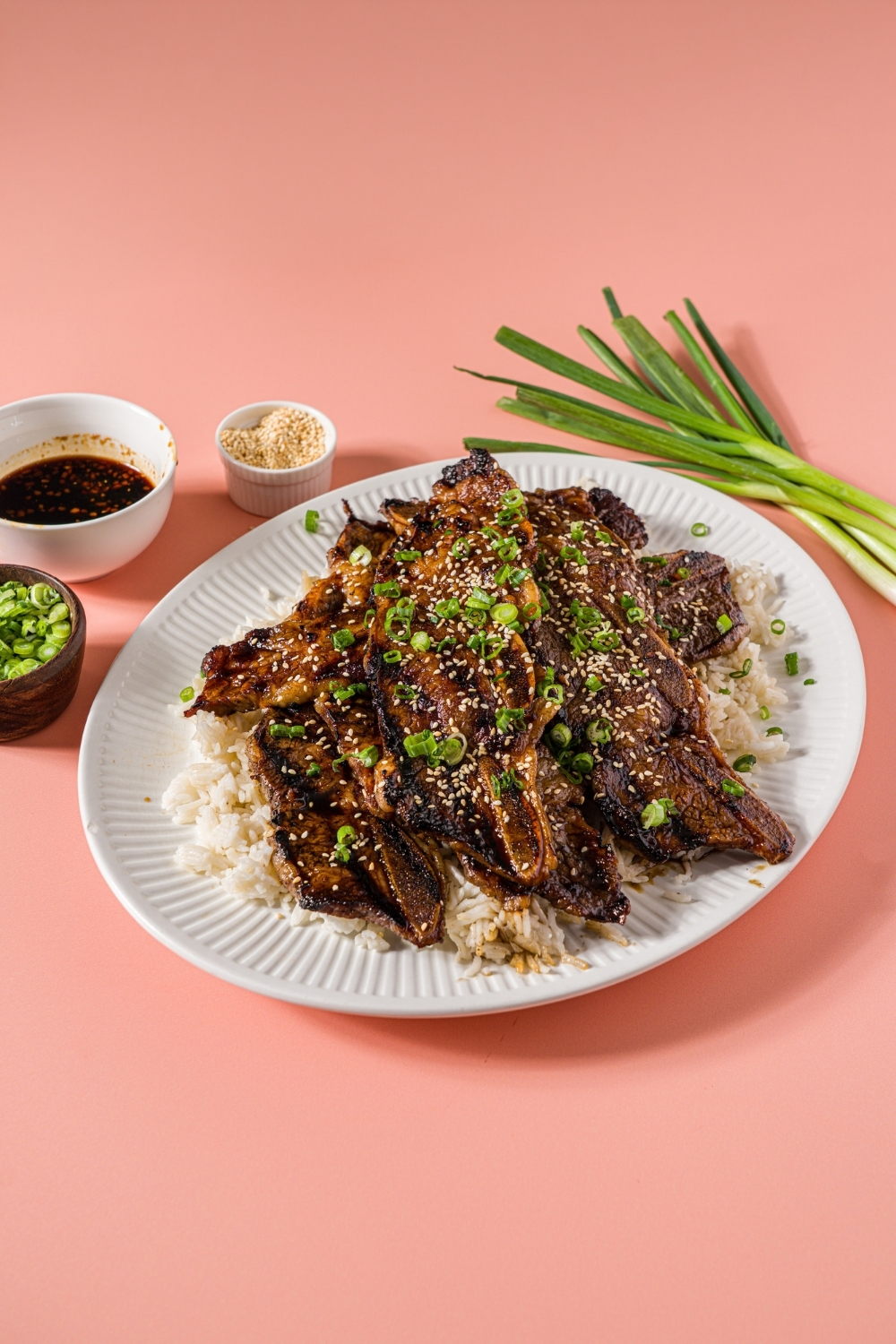 A white plate with flanken style short ribs served over white rice and garnished with sliced green onion and sesame seeds. The plate is on a pink counter with green onions and small bowls of garnishes.