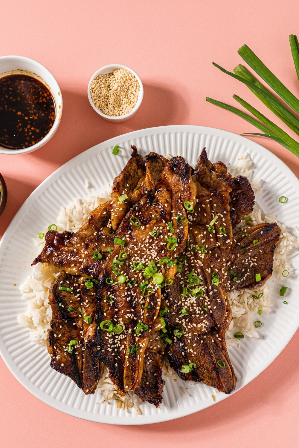 A white plate with flanken style short ribs served over white rice and garnished with sliced green onion and sesame seeds. The plate is on a pink counter with green onions and small bowls of garnishes.