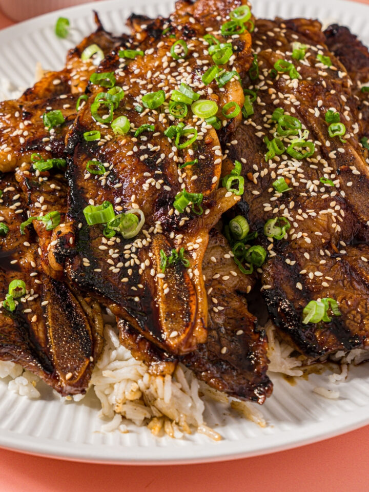 A white plate with flanken style short ribs served over white rice and garnished with sliced green onion and sesame seeds. The plate is on a pink counter.