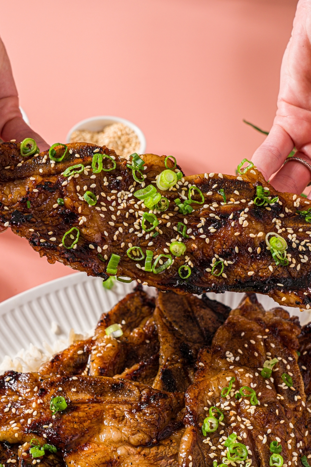 A white plate with flanken style short ribs served over white rice and garnished with sliced green onion and sesame seeds. A hand is holding one of the ribs. The plate is on a pink counter.