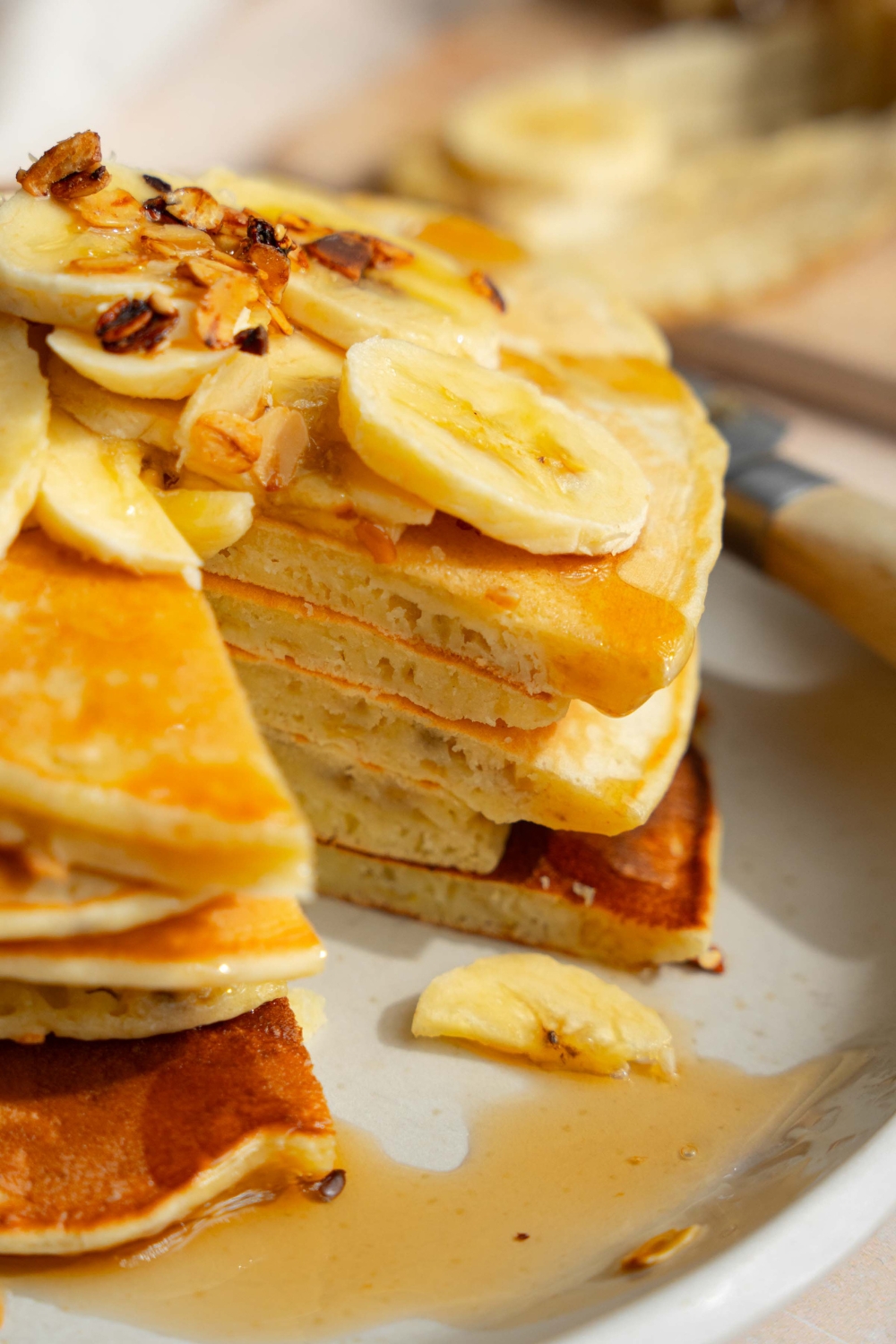 A white plate with a stack of fluffy banana pancakes topped with sliced banana and granola drizzled with syrup. There is a bite taken from the stack.