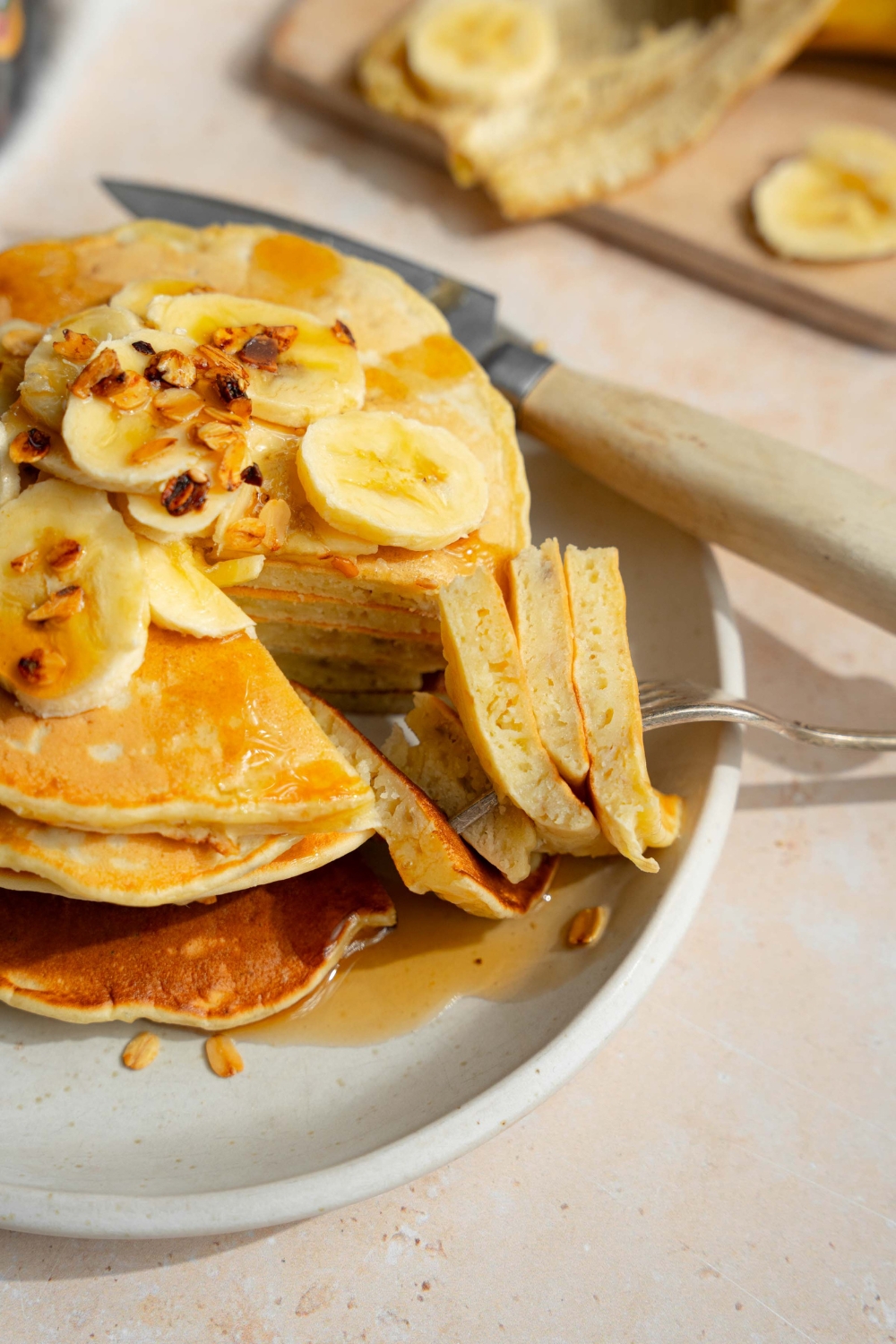 A white plate with a stack of fluffy banana pancakes topped with sliced banana and granola drizzled with syrup. A fork is taking a bite from the stack. The plate is on a tan counter with a knife and wooden cutting board with bananas.
