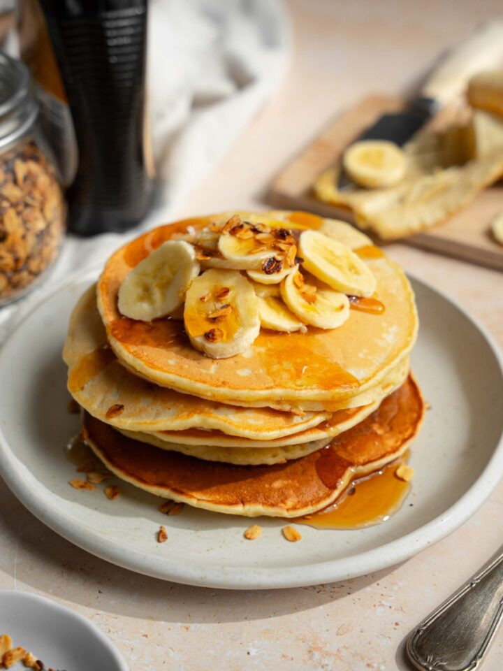 A white plate with a stack of fluffy banana pancakes topped with sliced banana and granola drizzled with syrup. The plate is on a tan counter with a bottle of syrup and wooden cutting board with bananas.