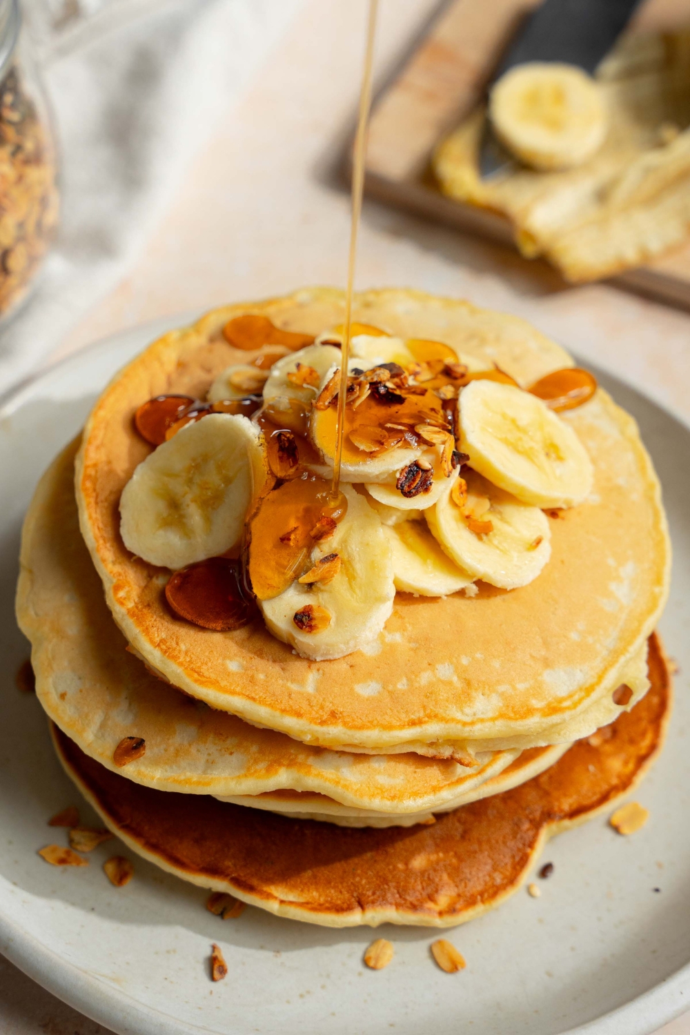A white plate with a stack of fluffy banana pancakes topped with sliced banana and granola. Syrup is being poured over the stack. The plate is on a tan counter with a bottle of syrup and wooden cutting board with bananas.