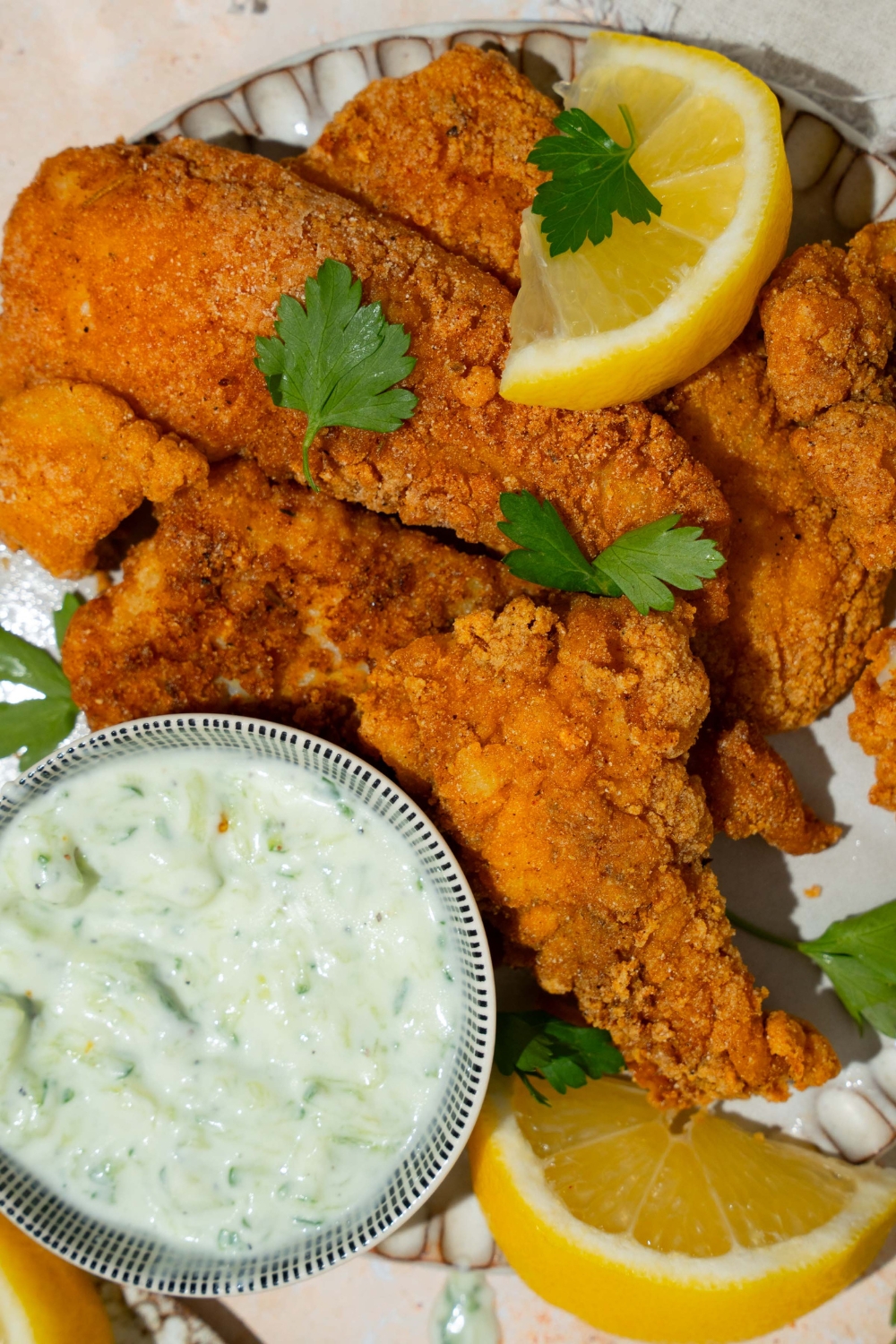 A white plate with several pieces of crispy Southern style fried fish garnished with fresh parsley and served with lemon and a small bowl of sauce. The plate is on a tan counter with a white cloth napkin.