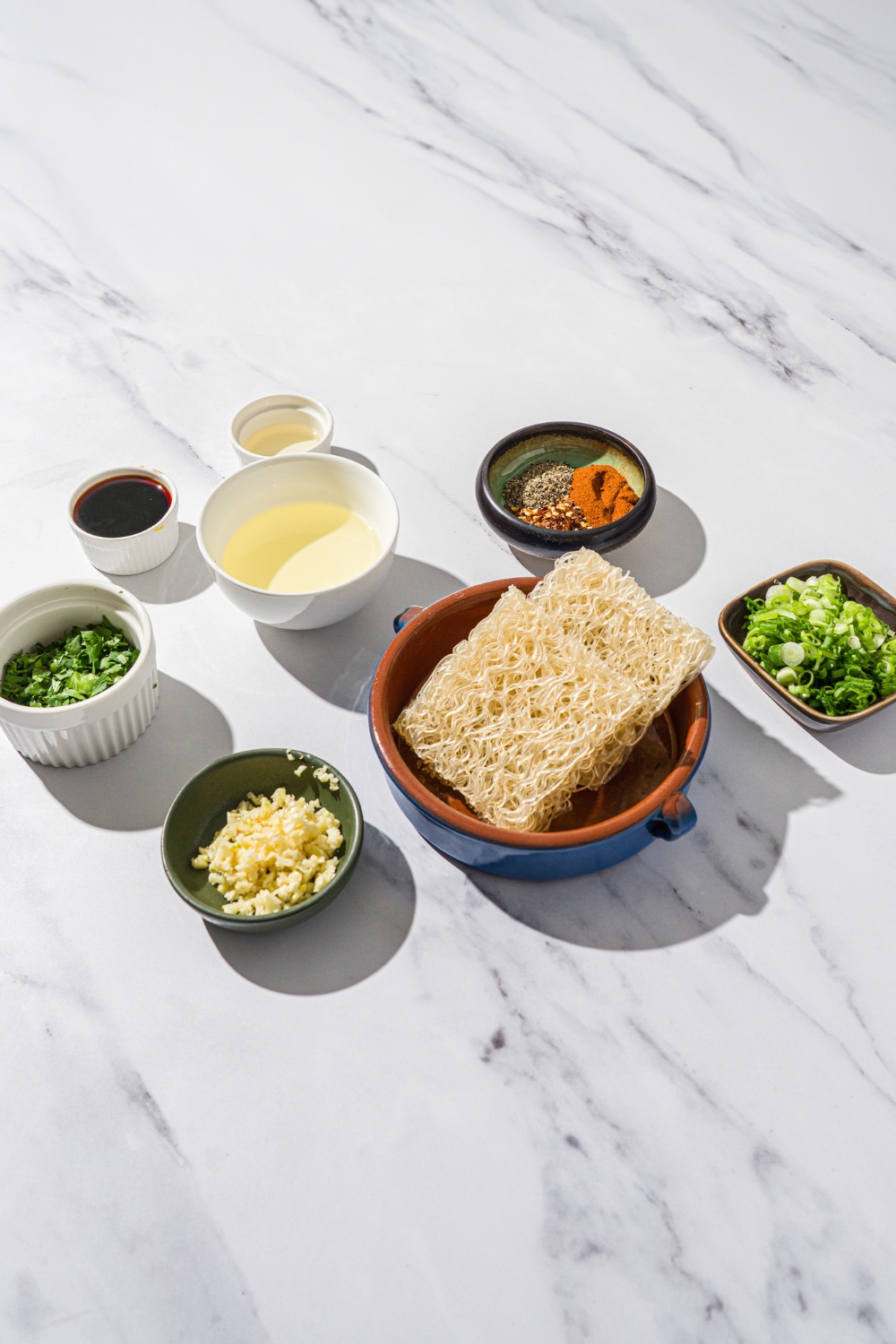 A marble counter with several bowls in various sizes containing ingredients to make garlic chili oil noodles including rice noodles, garlic, chili flakes, cayenne pepper, soy sauce, rice vinegar, green onions, oil, and seasonings.