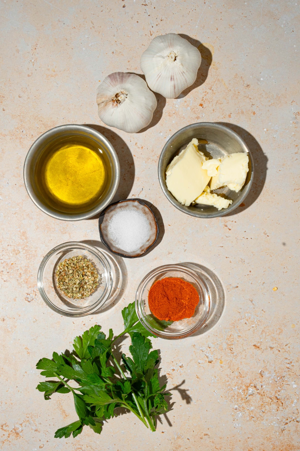 An overhead shot of several bowls in various sizes containing ingredients to make roasted garlic butter dip including garlic, butter, oil, and seasonings.