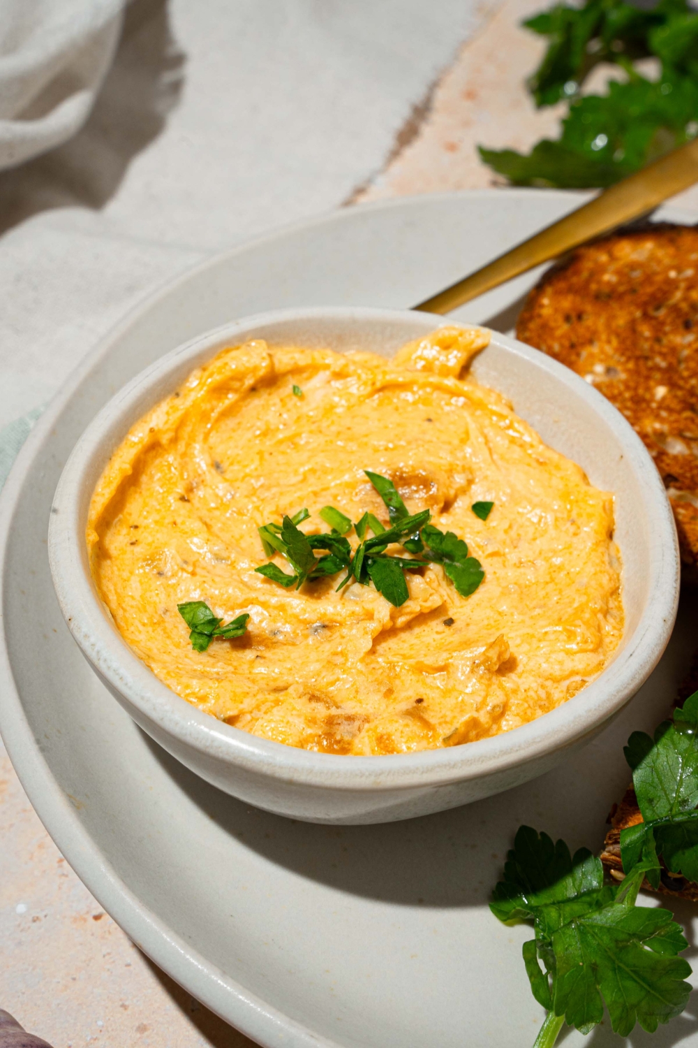 A white bowl with garlic dip garnished with fresh parsley. The bowl is on a white plate with a spoon and toasted bread. The plate is on a tan counter with a white cloth napkin.