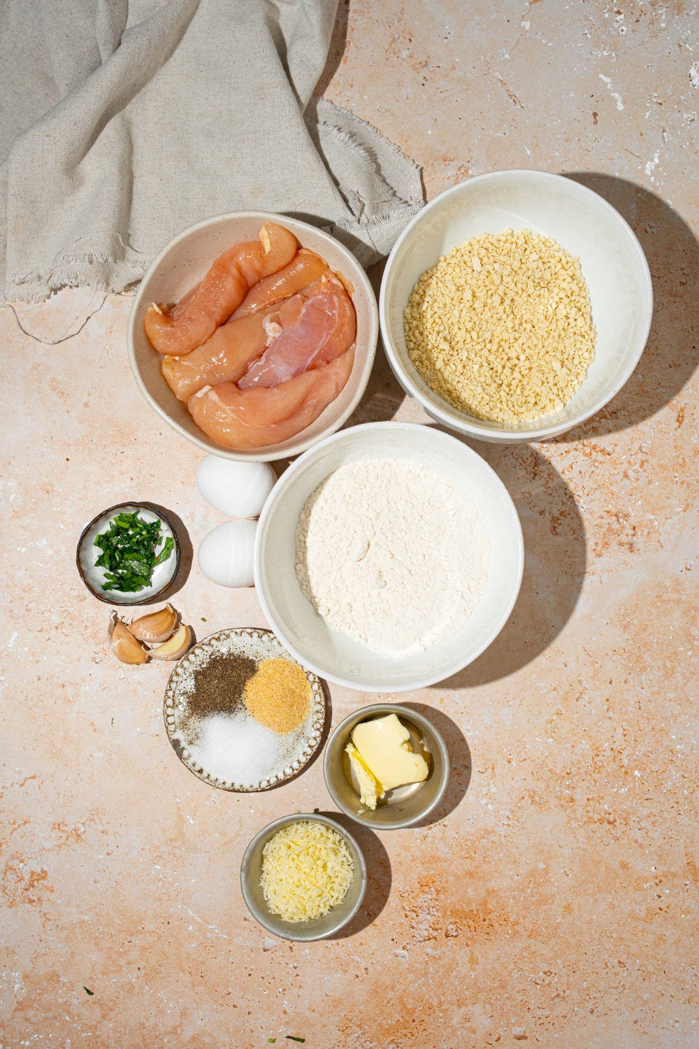 A tan counter with several bowls in various sizes containing ingredients to make garlic parmesan chicken tenders including chicken, flour, eggs, butter, panko breadcrumbs, parmesan cheese, and seasonings.