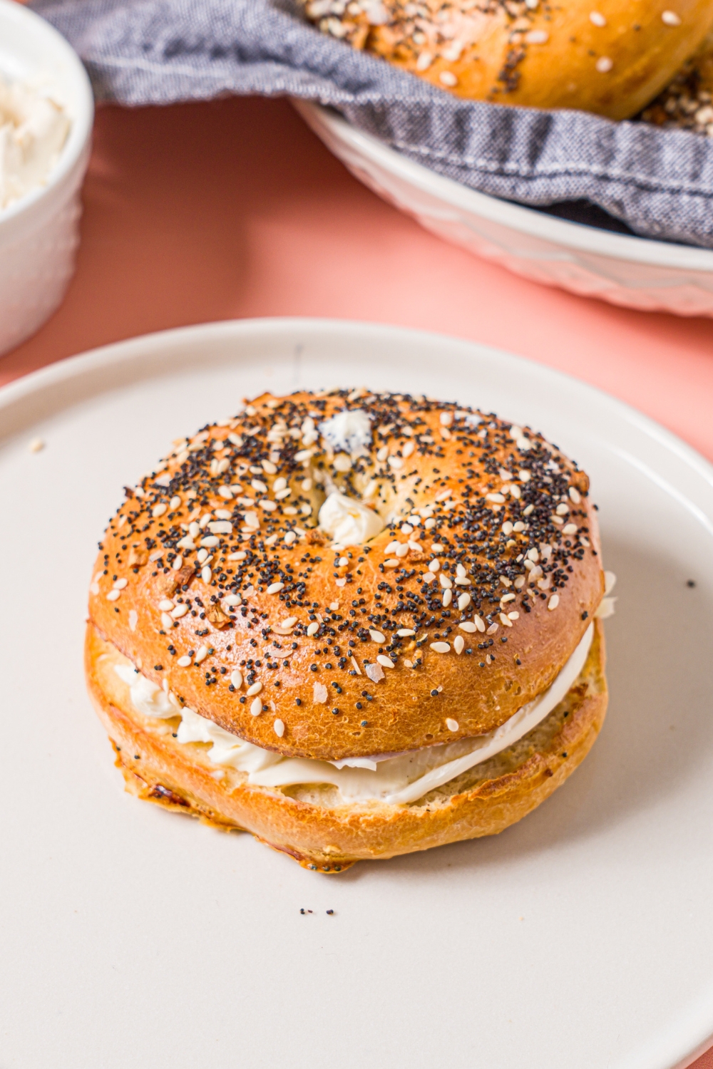 A white plate with a whole greek yogurt everything bagel with cream cheese. The plate is on a pink counter with a bowl of bagels.