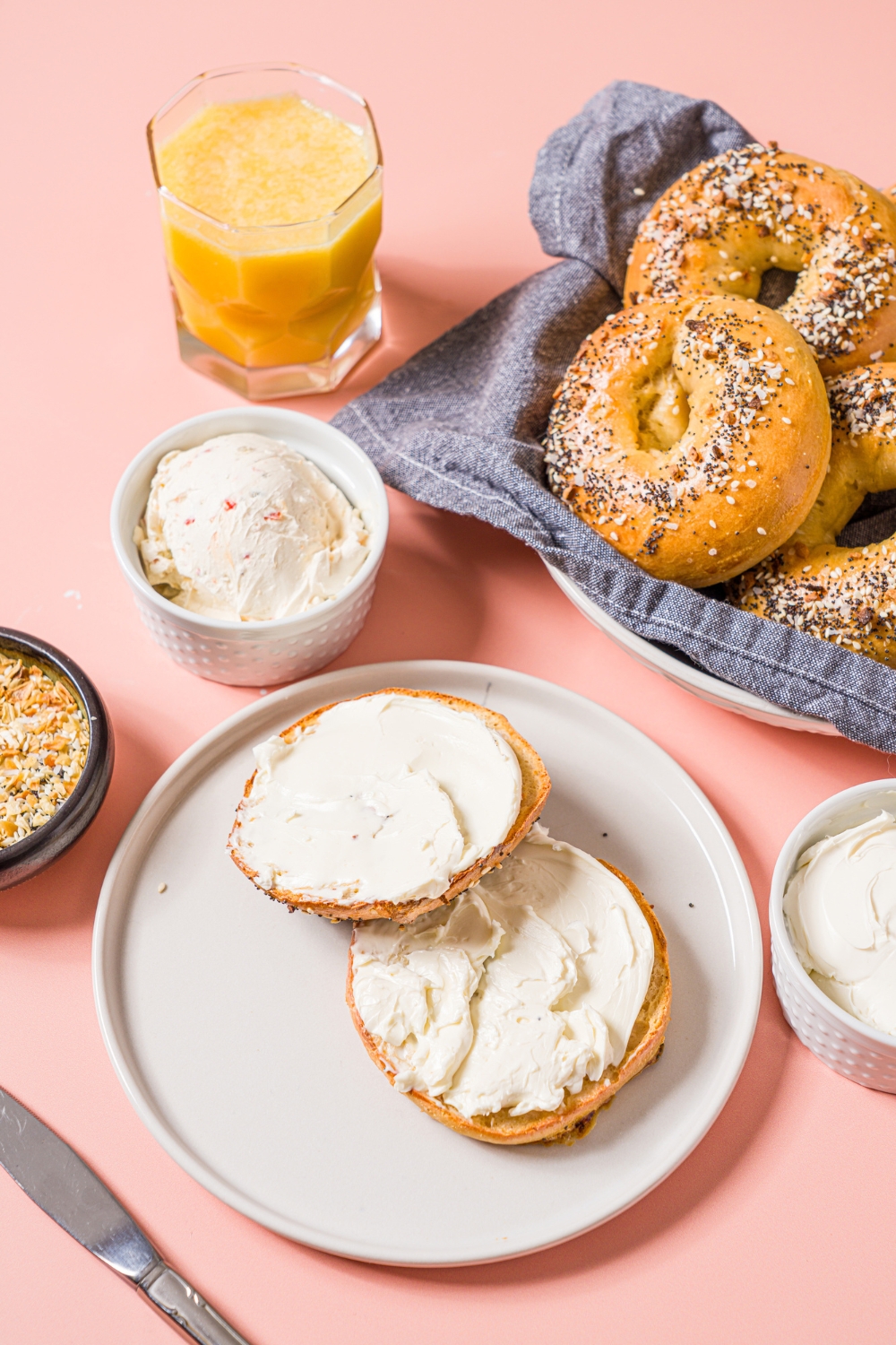 A bowl of greek yogurt everything bagels with a blue cloth napkin. The bowl is on a pink counter with a plate of a sliced bagel with cream cheese, small bowl of cream cheese, and orange juice.
