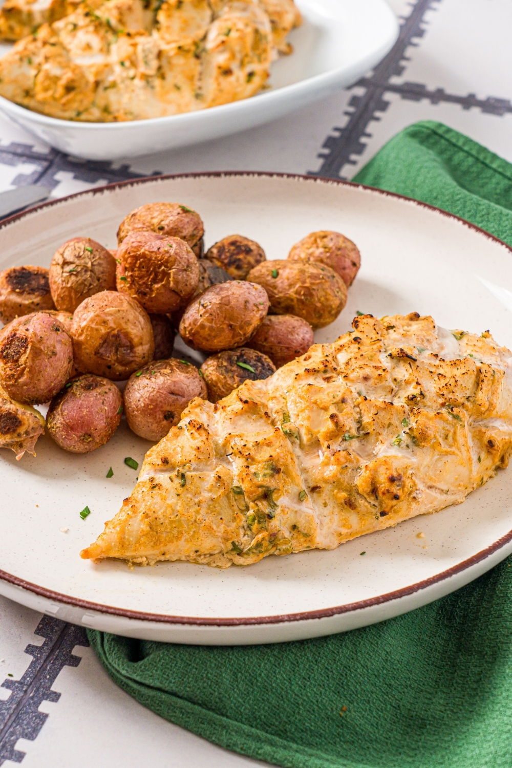 A white plate with greek yogurt marinated chicken garnished with fresh parsley and served with baked potatoes. The plate is on a tiled counter with a green cloth napkin and an additional bowl of chicken.