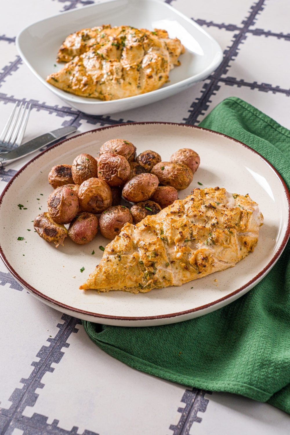 A white plate with greek yogurt marinated chicken garnished with fresh parsley and served with baked potatoes. The plate is on a tiled counter with a green cloth napkin and an additional bowl of chicken.