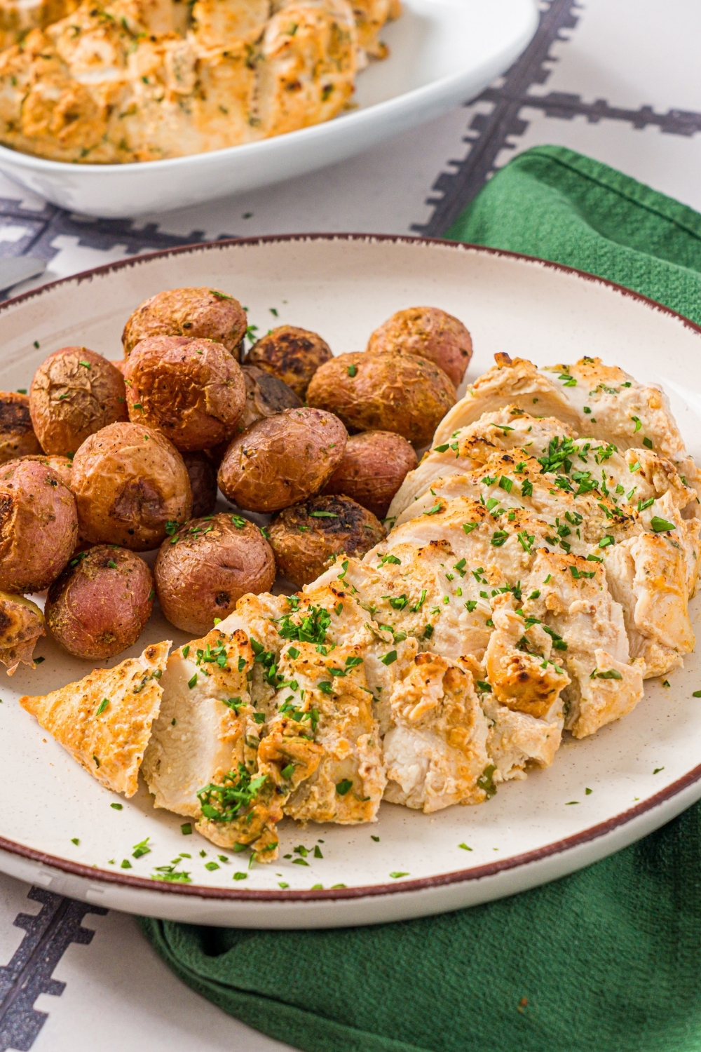 A white plate with sliced greek yogurt marinated chicken garnished with fresh parsley and served with baked potatoes. The plate is on a tiled counter with a green cloth napkin and an additional bowl of chicken.