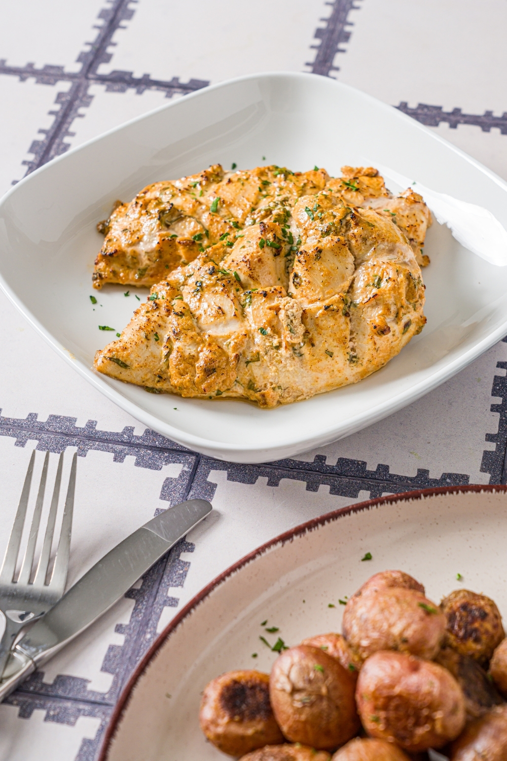 A white plate with sliced greek yogurt marinated chicken garnished with fresh parsley. The plate is on a tiled counter with a plate of potatoes.