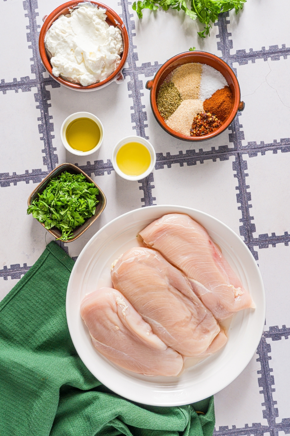 A tiled counter with several bowls in various sizes containing ingredients to make greek yogurt marinated chicken including chicken breasts, greek yogurt, oil, lemon, fresh parsley, and seasonings.