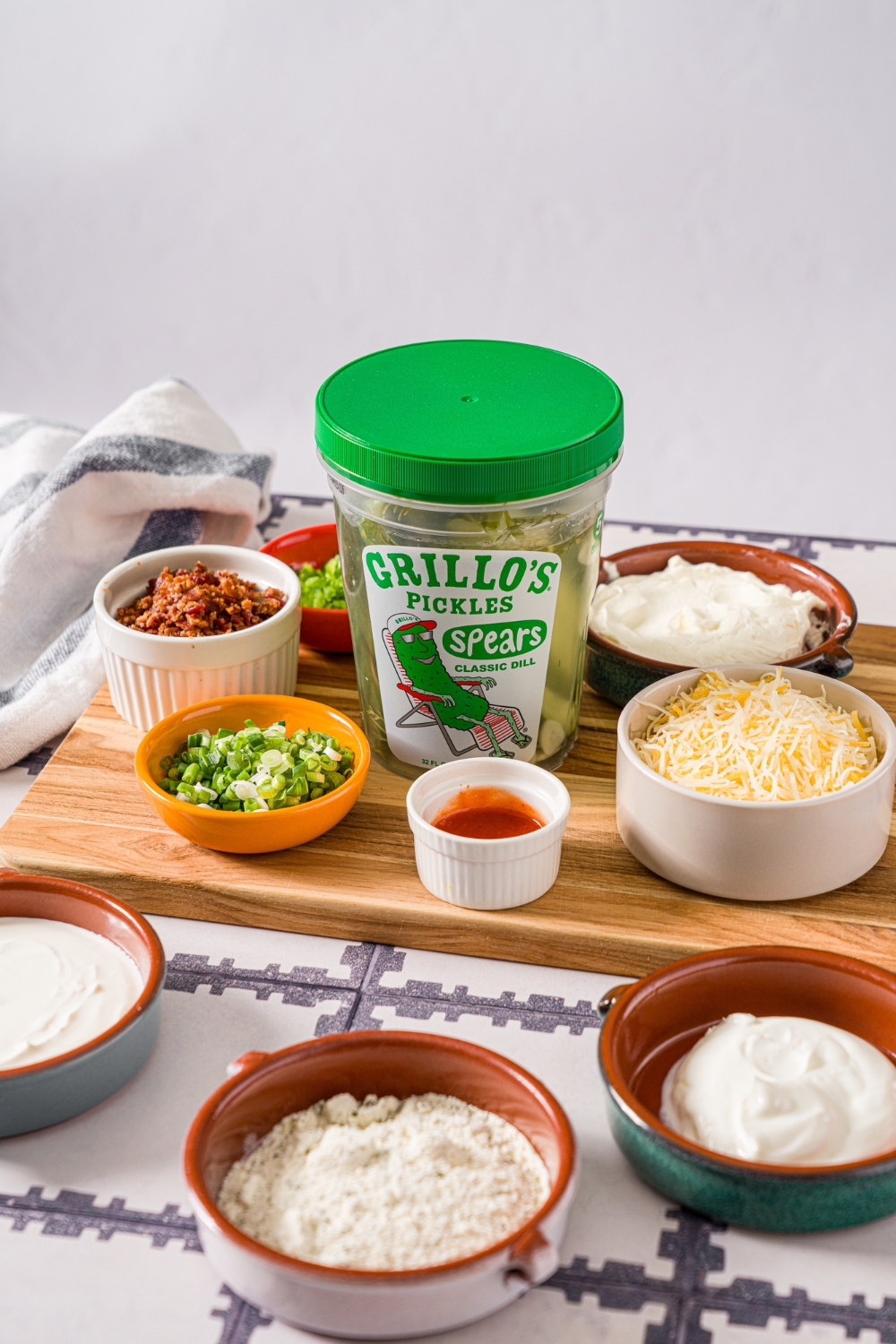A tub of Grillo's pickles, a bowl of cream cheese, a bowl of Greek yogurt, a bowl of sour cream, a bowl of jalapeños, a bowl of shredded cheddar cheese, and a bowl of bacon.
