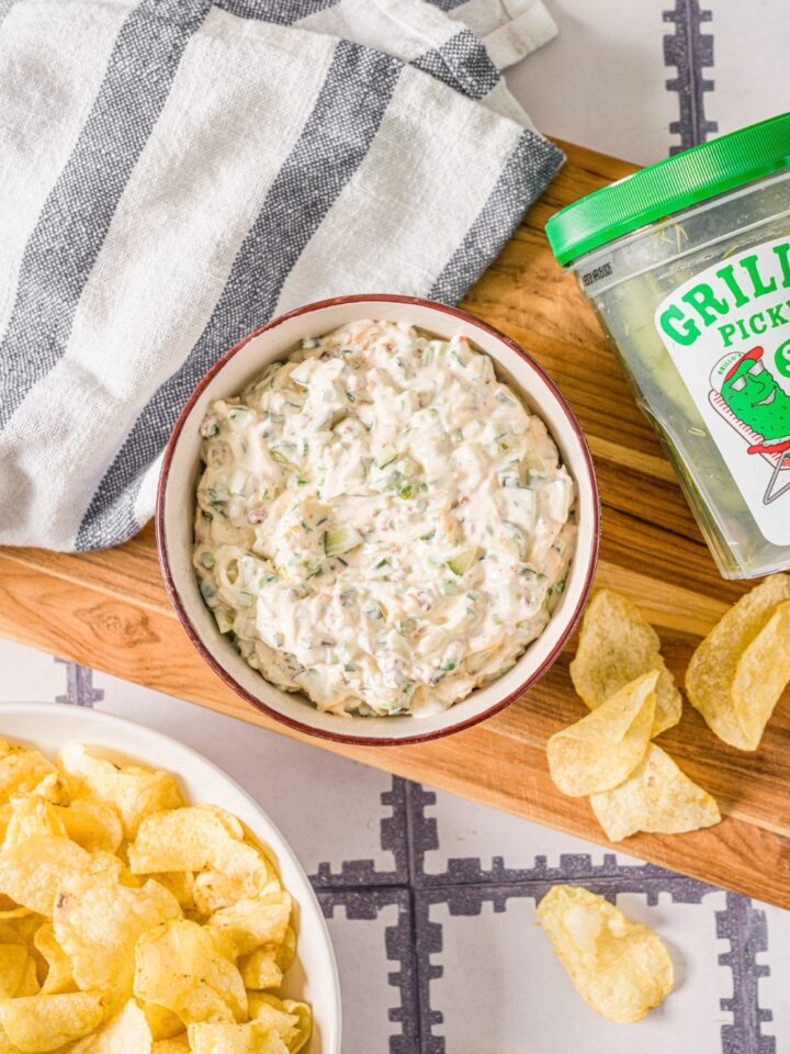 A creamy grilled pickle dip in a white bowl on top of a wooden cutting board with a tub of Grillo's pickles next to the dip and some potato chips on the cutting board as well.